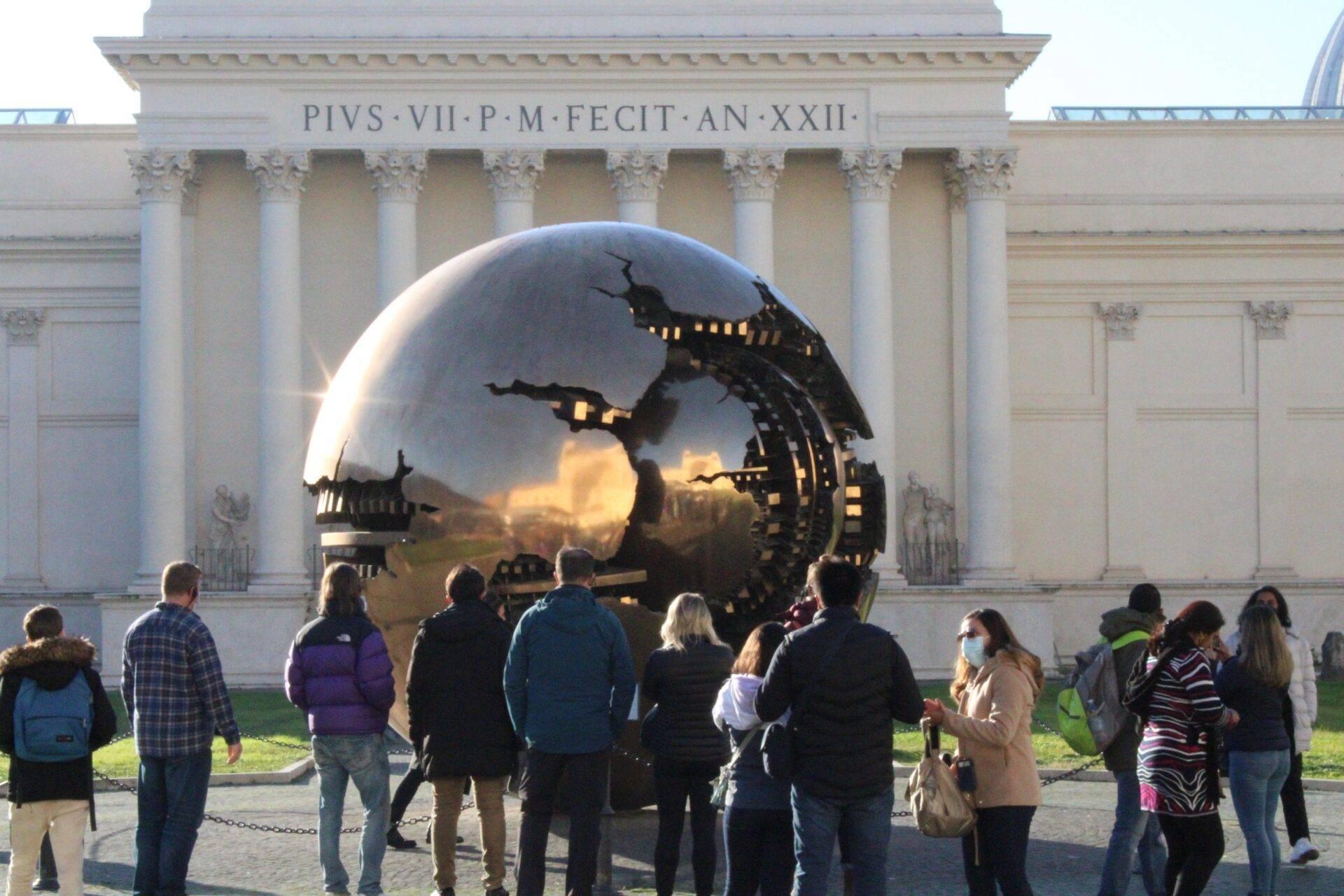 A group of visitors admiring a giant reflective metal sphere in front of a classical building with white columns