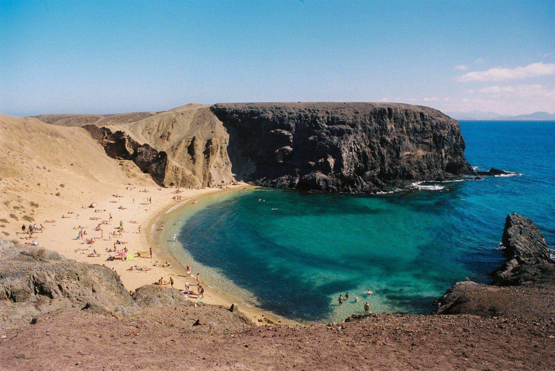 Una spiaggia di sabbia dorata incorniciata da scogliere nere e un mare turchese cristallino con bagnanti