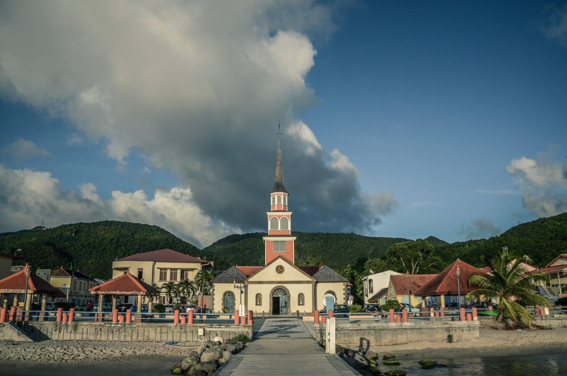Bunte Kirche am Meer mit Kirchturm, umgeben von rotbedachten Häusern und grünen Bergen