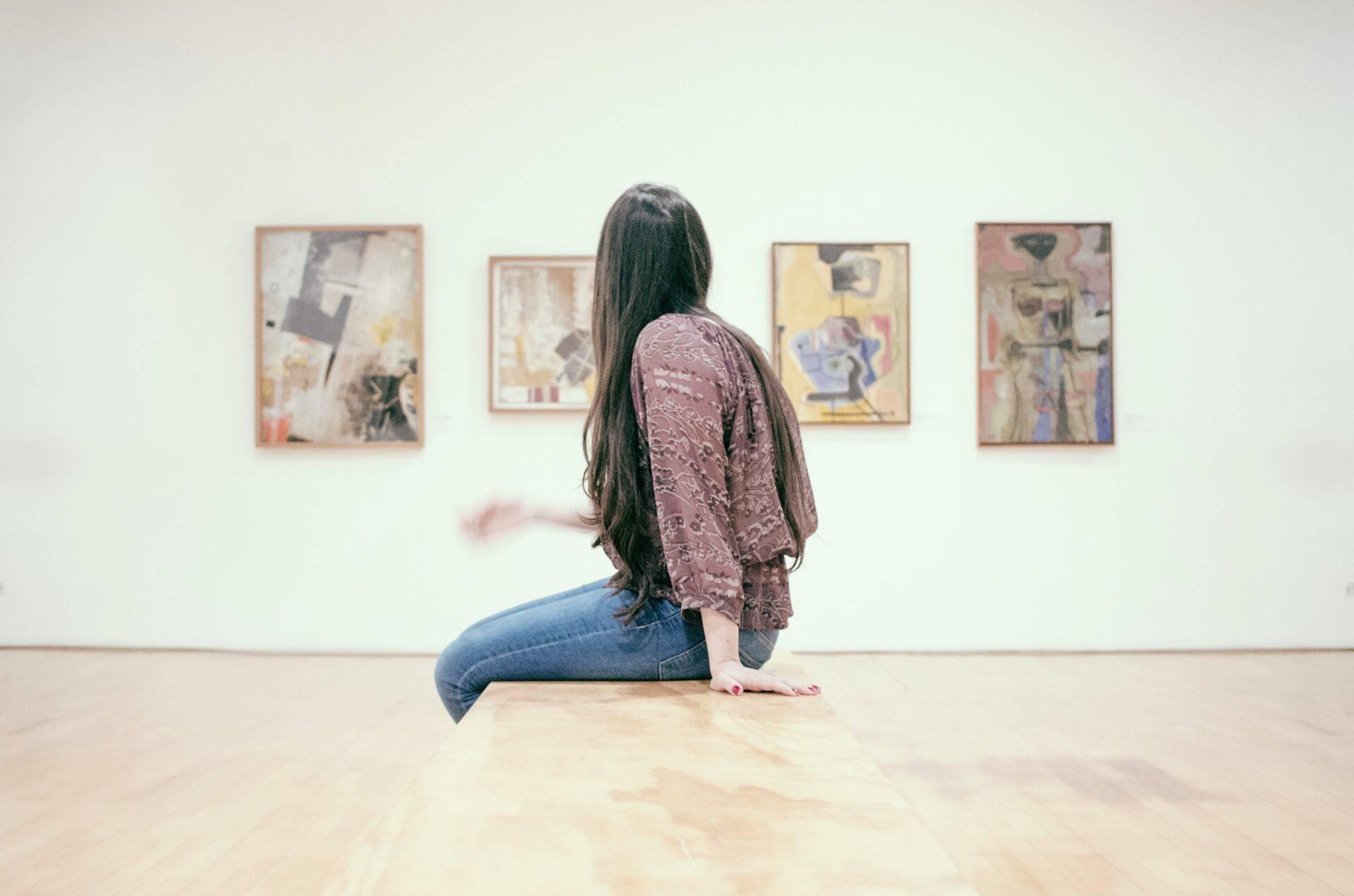 Eine Frau sitzt und betrachtet abstrakte Gemälde in einer modernen Kunstgalerie mit weißen Wänden