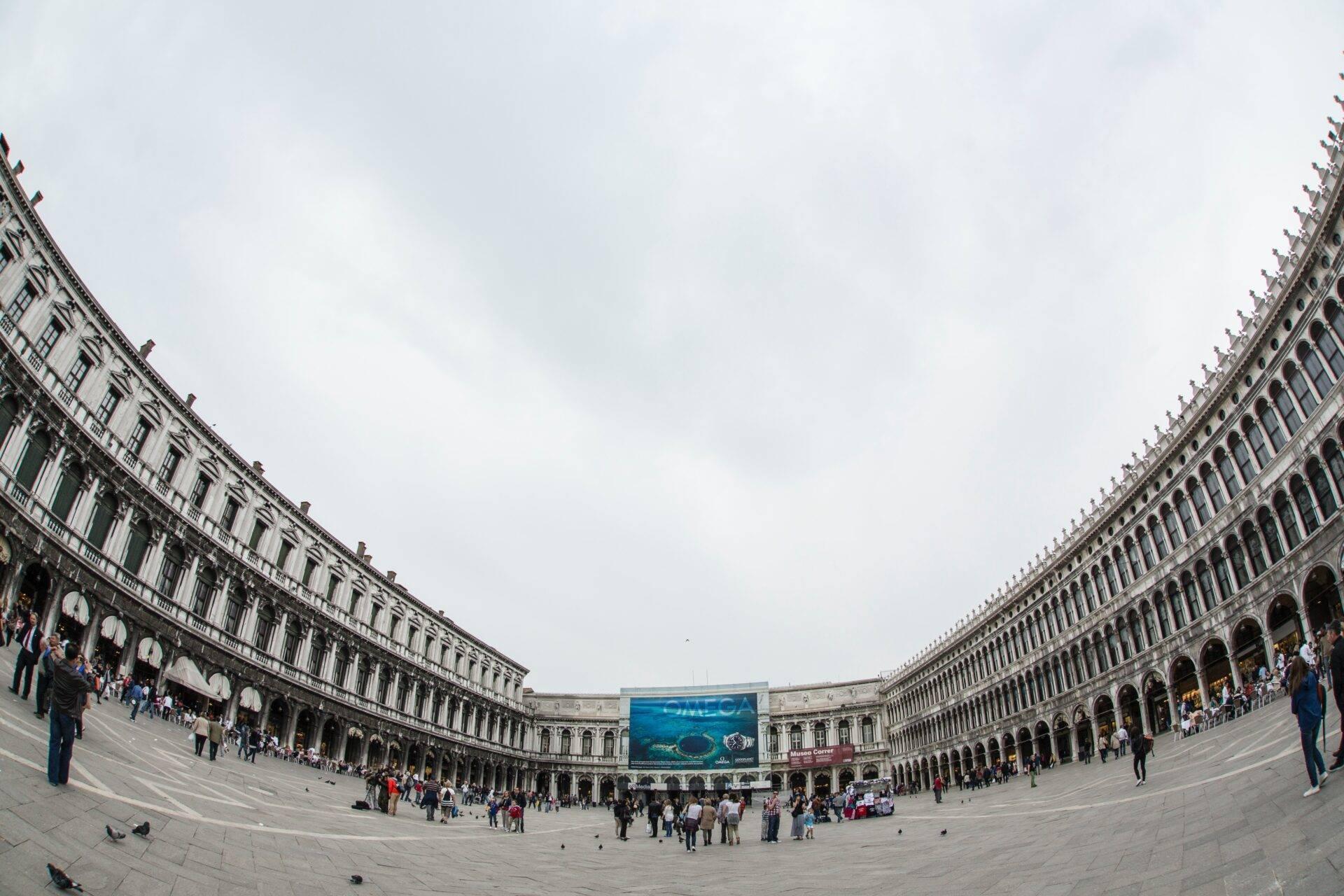 A large square ringed by elegant historic arcaded buildings, filled with tourists