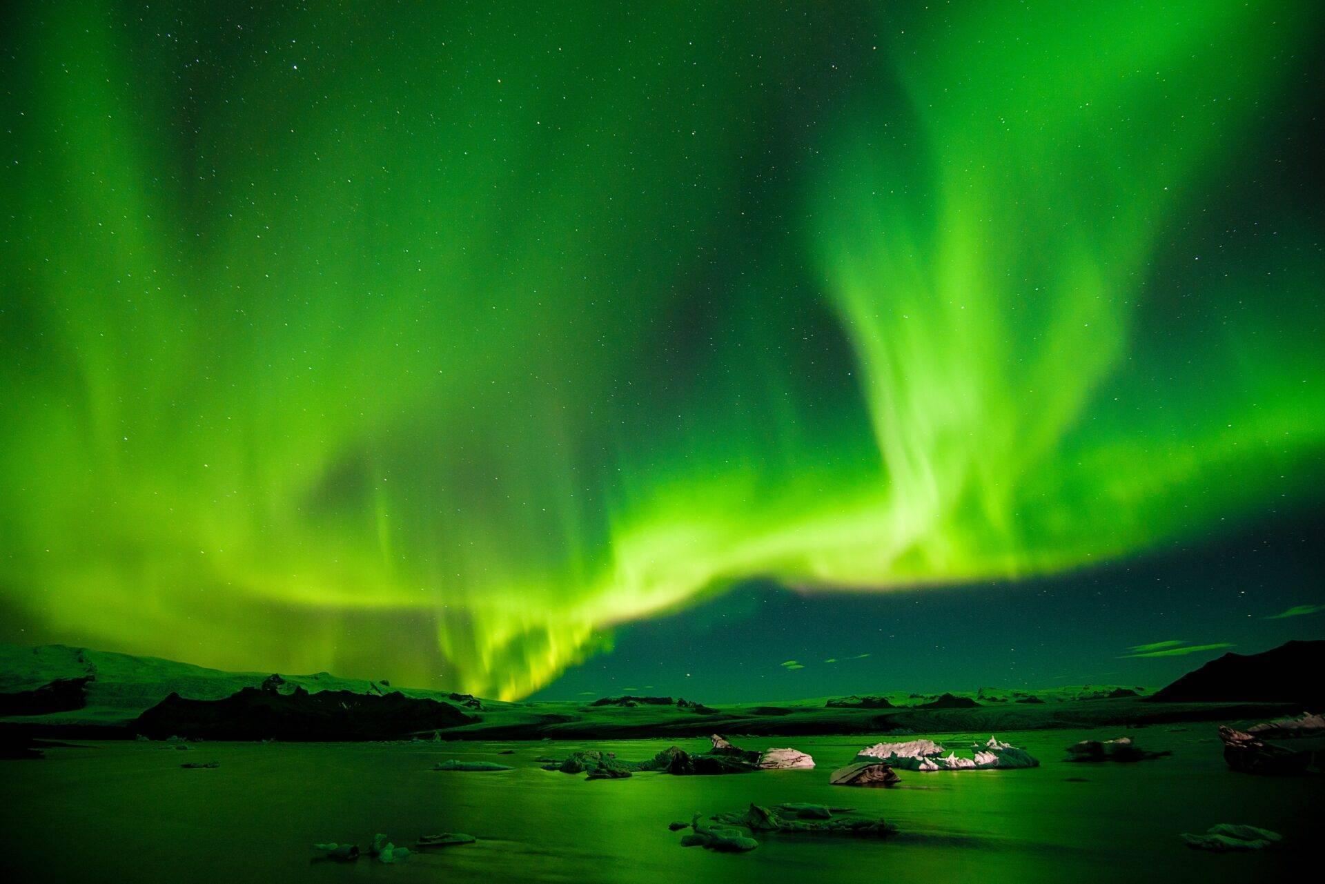 Aurora boreal verde y espectacular danzando sobre un paisaje helado con rocas y montañas al fondo