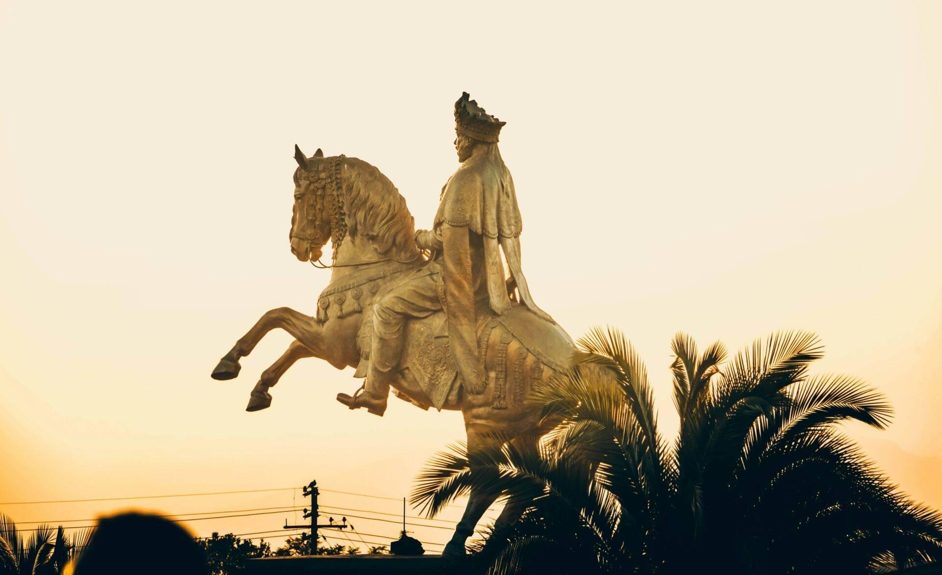 Golden equestrian statue of a rider in royal dress at sunset, surrounded by palm trees