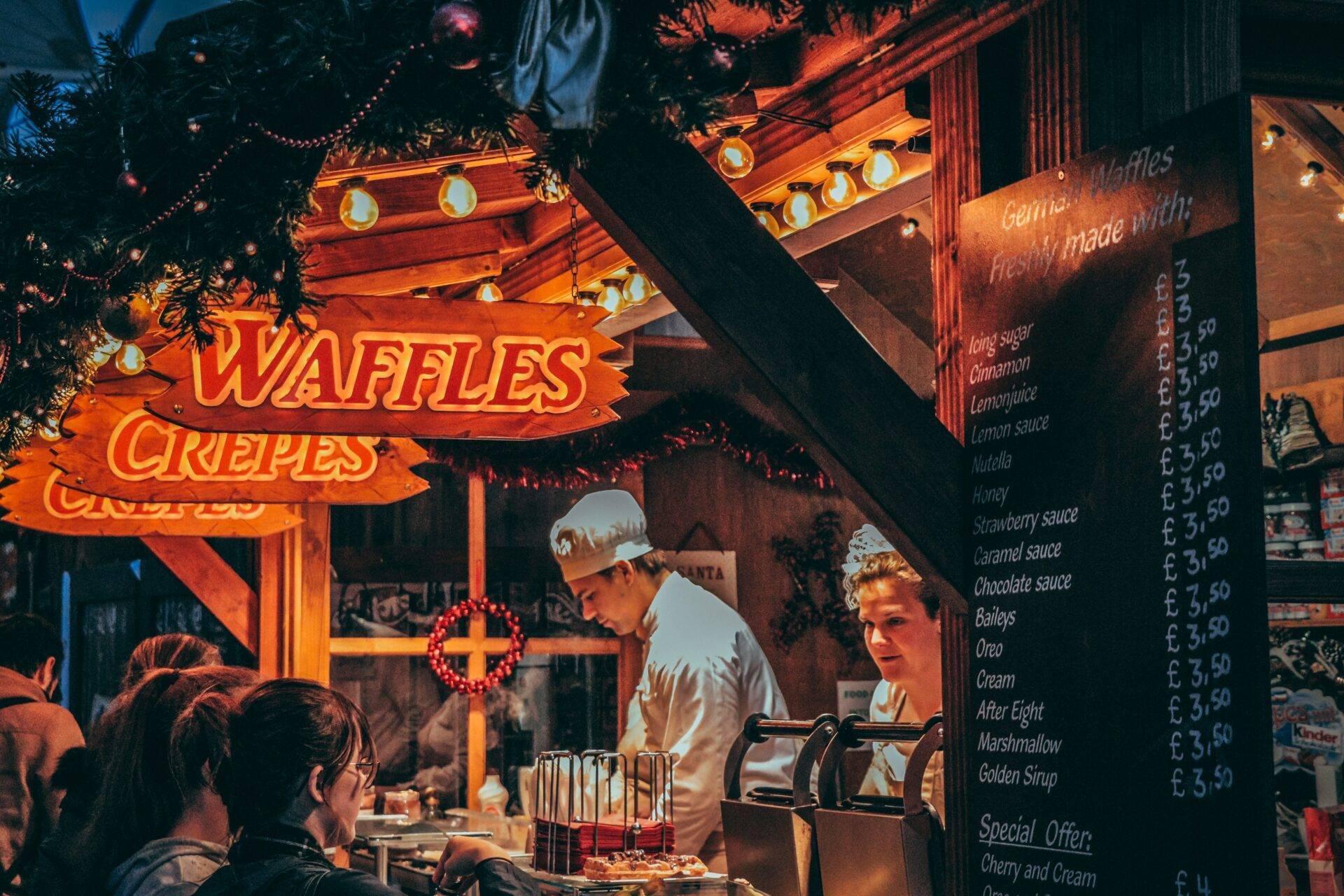 Un puesto de crepes y gofres iluminado en un mercado navideño europeo con el menú expuesto y vendedores en acción