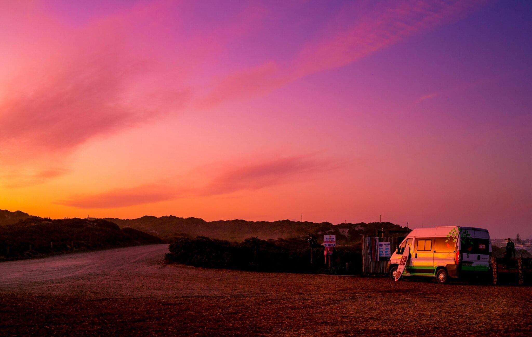 Beleuchtetes Wohnmobil beim Sonnenuntergang in einer Wüstenlandschaft mit rosafarbenem und orangefarbenem Abendhimmel