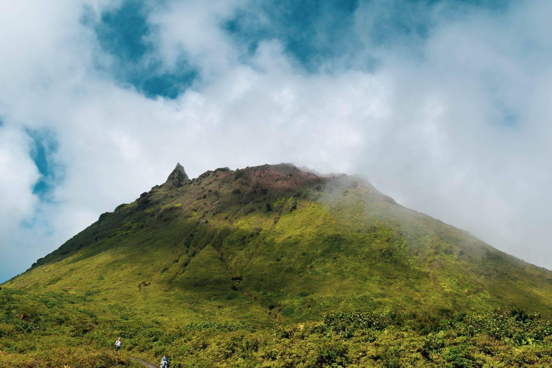 Ein konischer Vulkan mit üppiger Vegetation und teils wolkenverhülltem Gipfel unter blauem Himmel