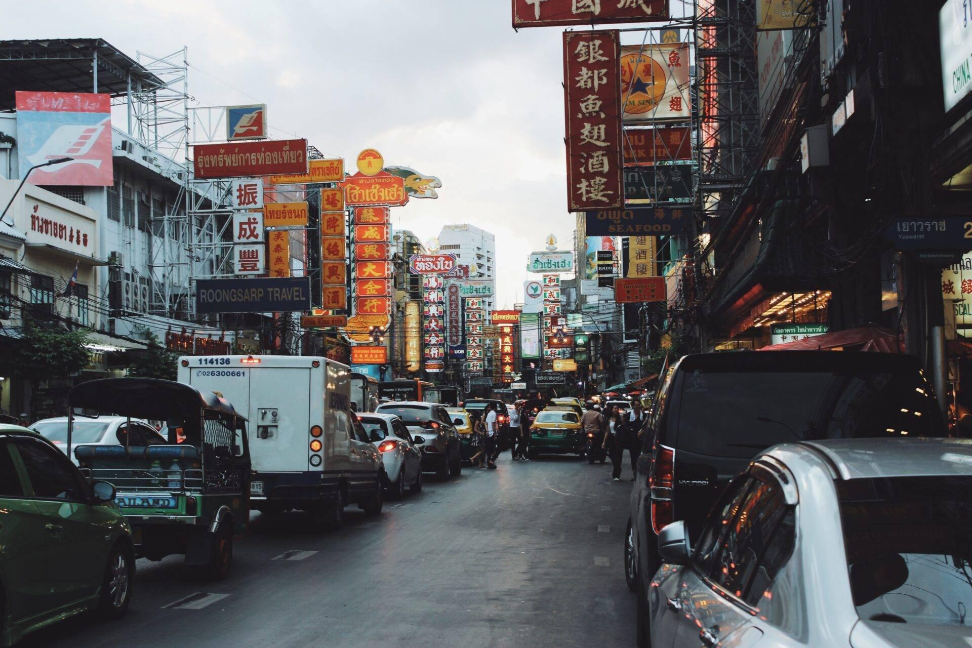 Una vivace strada commerciale di Bangkok con insegne cinesi colorate, auto e pedoni sotto un cielo nuvoloso