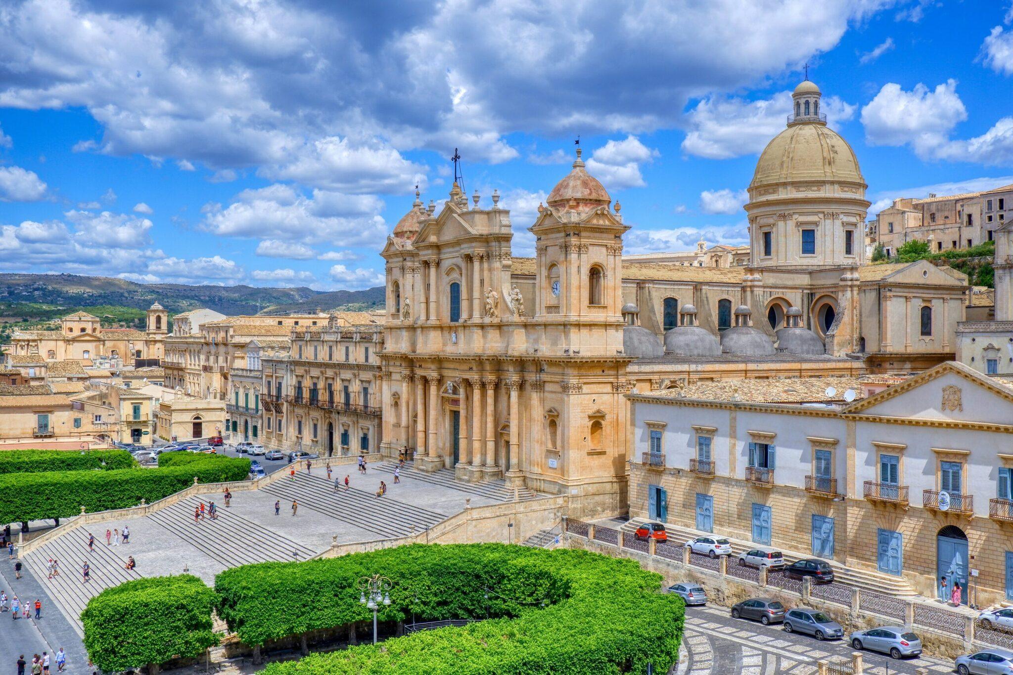 Una magnífica catedral barroca con cúpula dorada domina una piazza empedrada en una ciudad histórica siciliana bajo un cielo azul