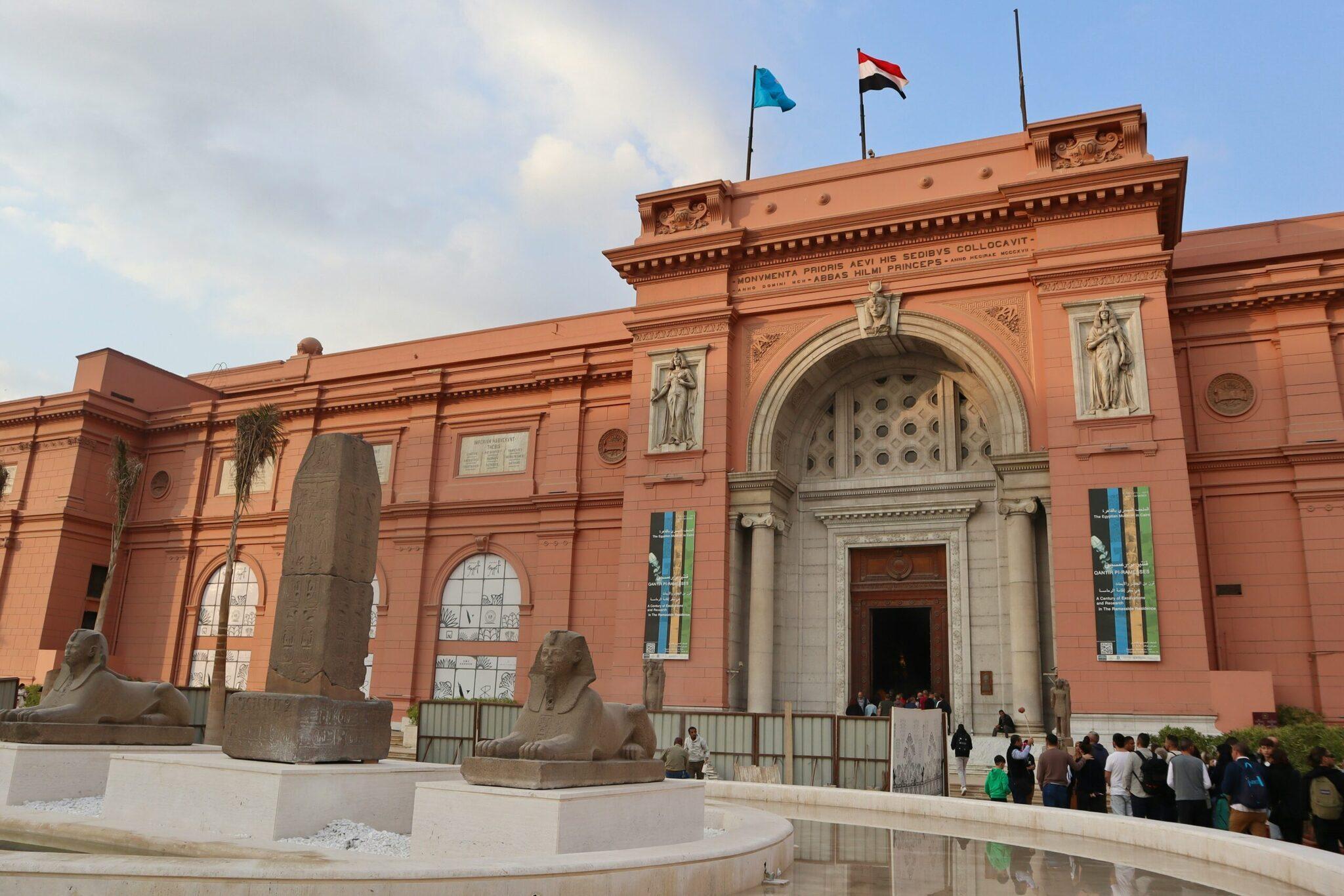 Fachada rosa de un museo egipcio con estatuas de esfinge, obelisco y banderas, con visitantes en la entrada