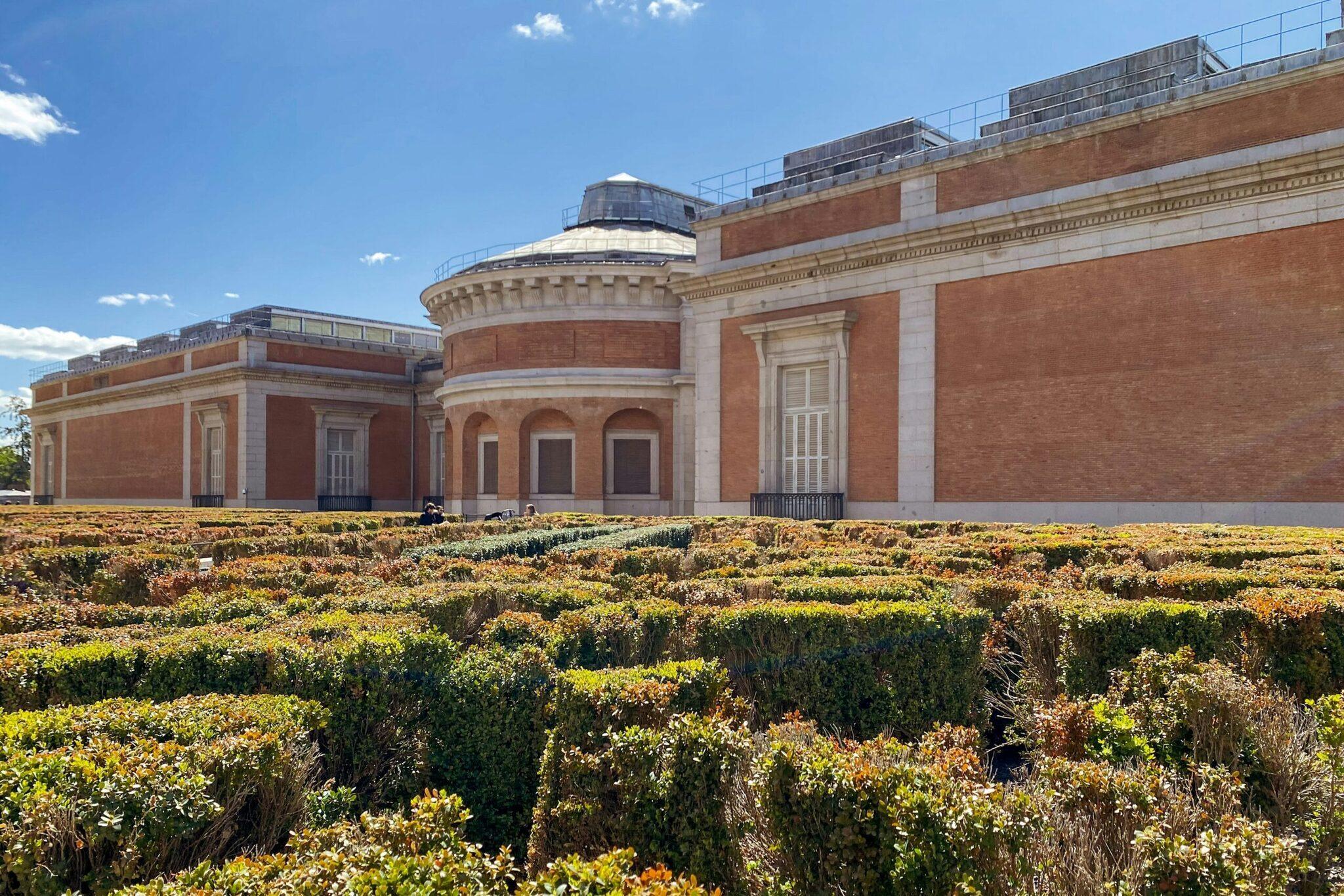 Museo con facciata in mattoni rossi e cupola, circondato da un giardino topiario geometrico sotto un cielo blu