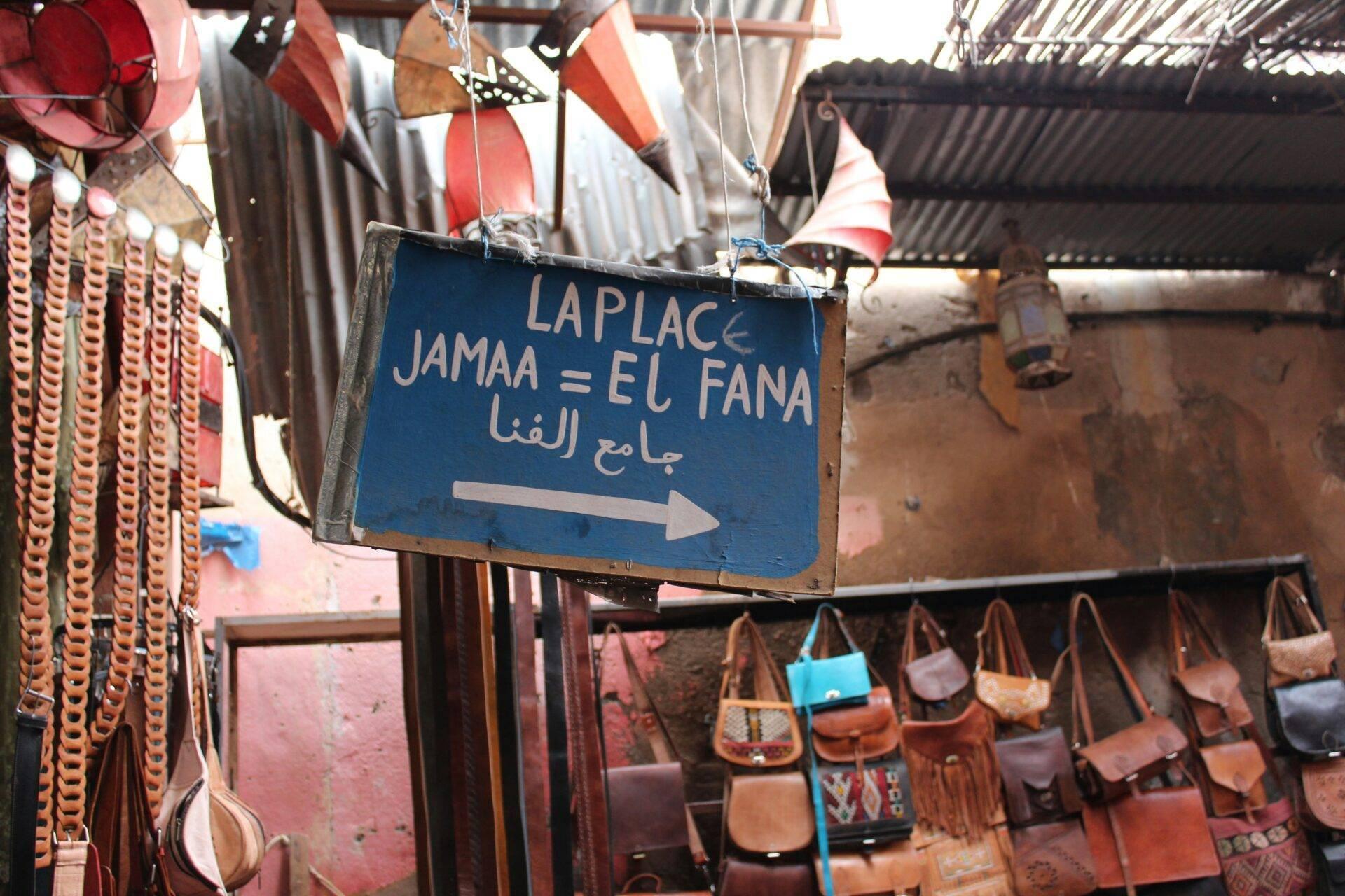 Cartel indicador azul en francés y árabe señalando una tienda de marroquinería con bolsos de cuero expuestos en una medina marroquí