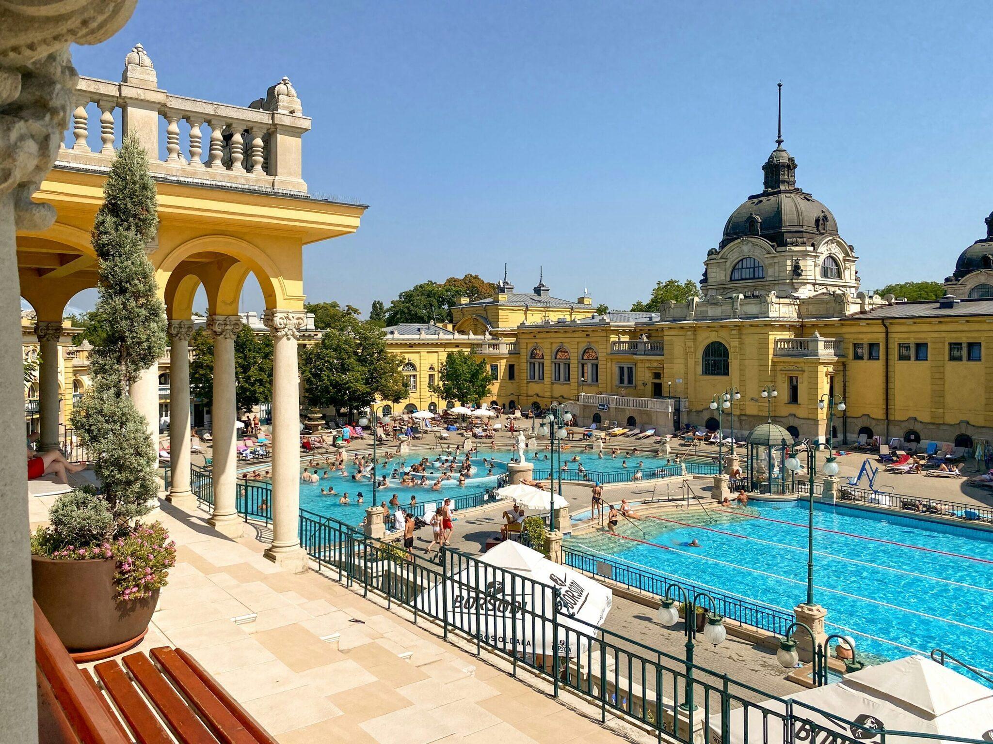 Panoramic view of the thermal baths with turquoise pools, yellow neo-Baroque architecture and a grand dome, bathers relaxing in fine weather