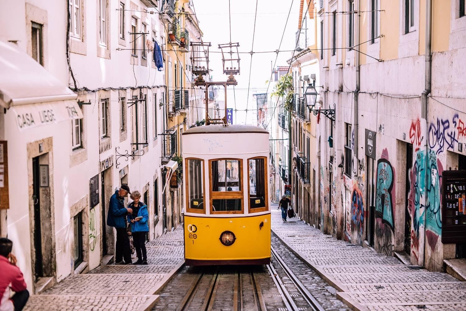 Eine historische gelbe Straßenbahn fährt eine gepflasterte, schmale Gasse hinauf, gesäumt von bunten Gebäuden einer europäischen Stadt