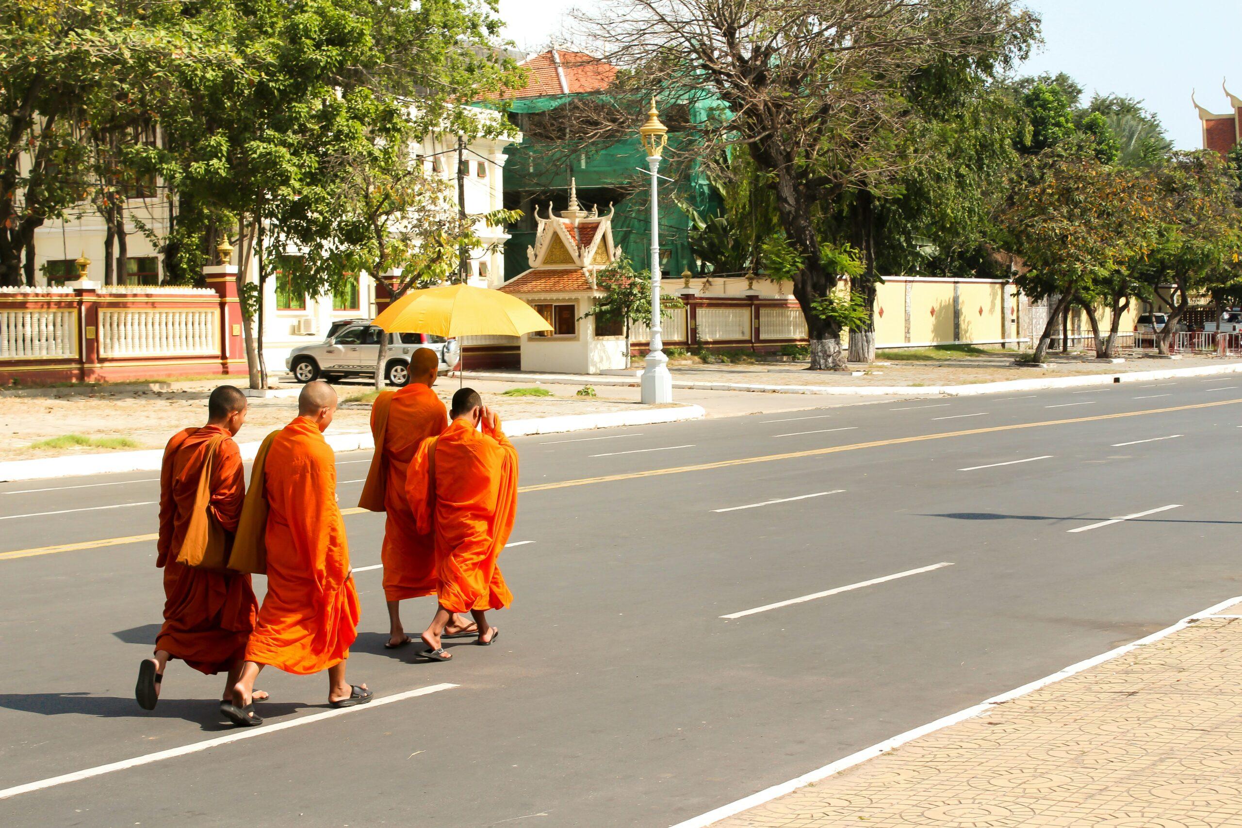 Vier Mönche in orangefarbenen Roben überqueren eine breite, sonnenbeschienene Allee vor einem traditionellen buddhistischen Tempel