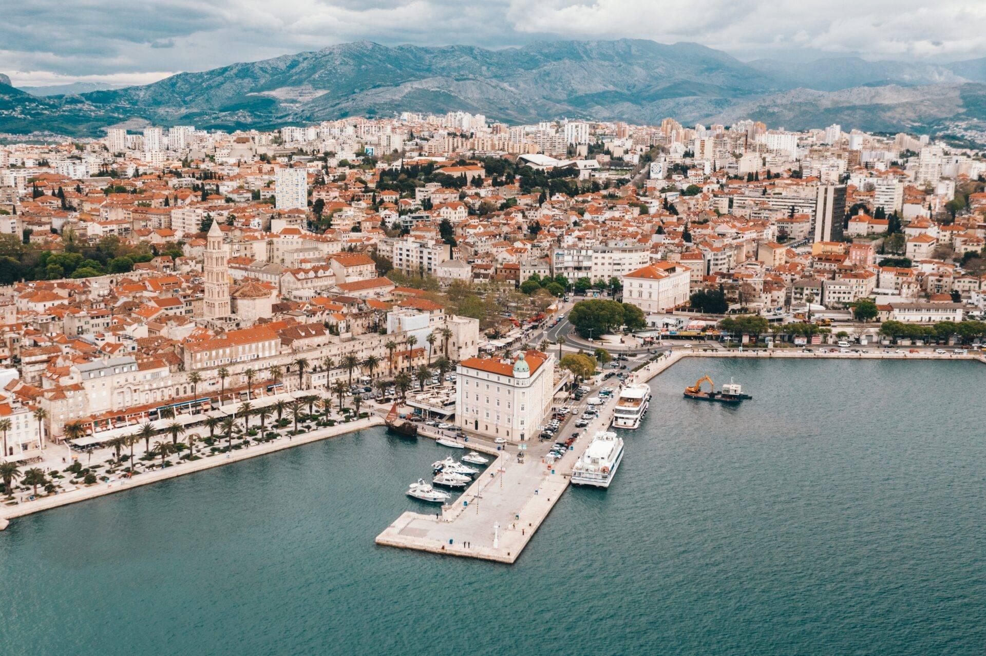 Veduta aerea di una città costiera mediterranea con porto, tetti color terracotta e montagne verdeggianti sullo sfondo