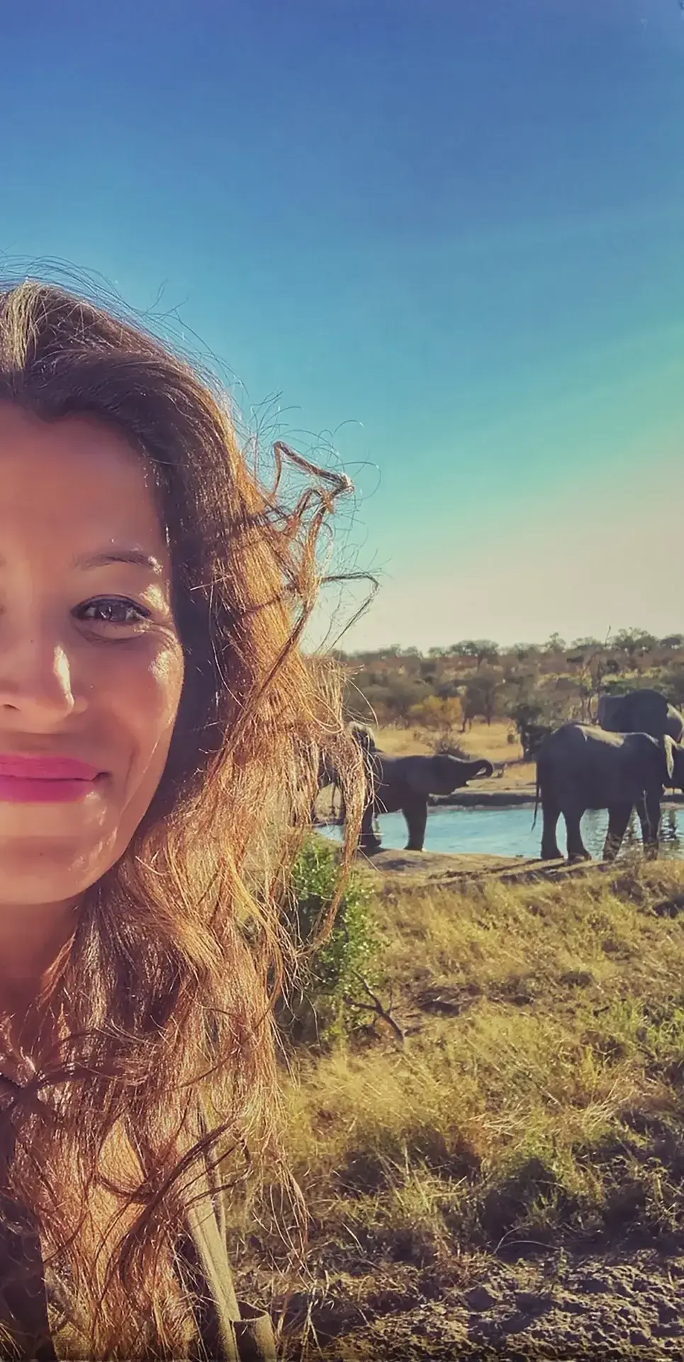 Selfie de una mujer sonriente con una manada de elefantes en un abrevadero de la sabana africana