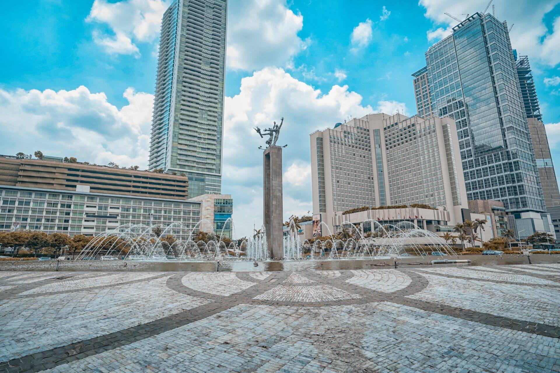 Piazza urbana lastricata con una statua monumentale, fontane zampillanti e grattacieli moderni sotto un cielo azzurro