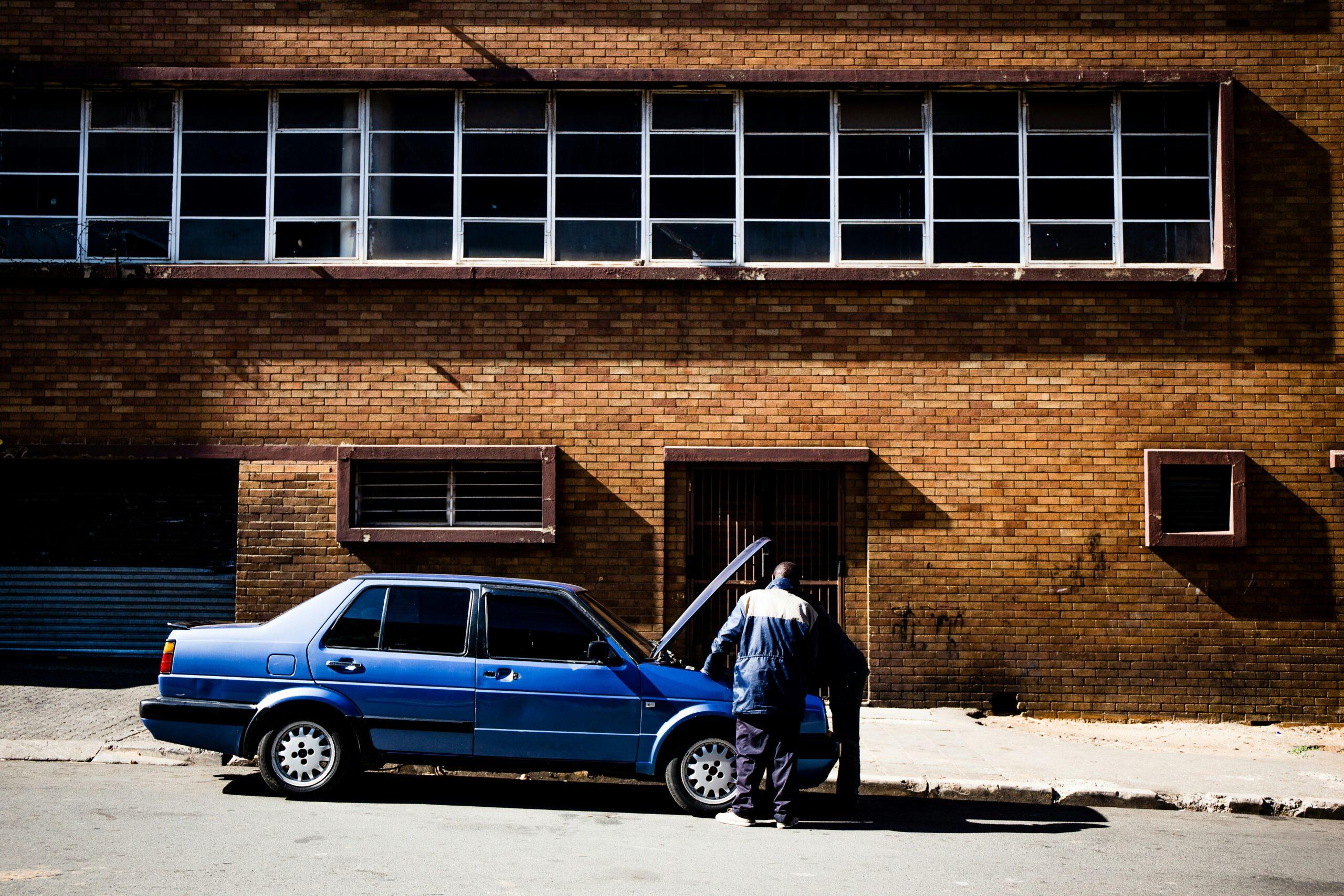 Un mecánico repara un coche azul frente a un edificio industrial de ladrillo con ventanas enrejadas