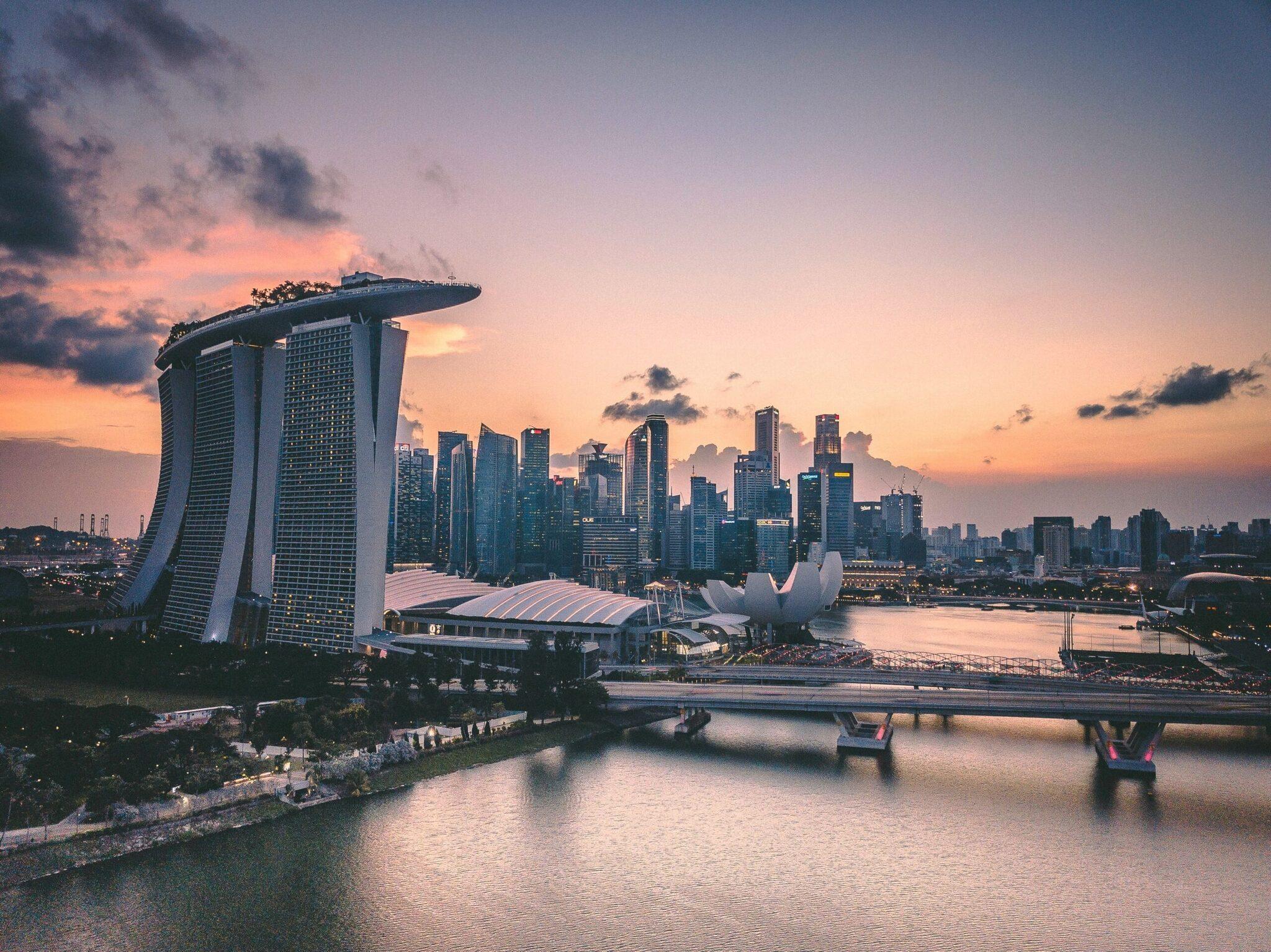 Skyline urbano al tramonto con grattacieli moderni e un futuristico edificio a forma di vela affacciato sull’acqua