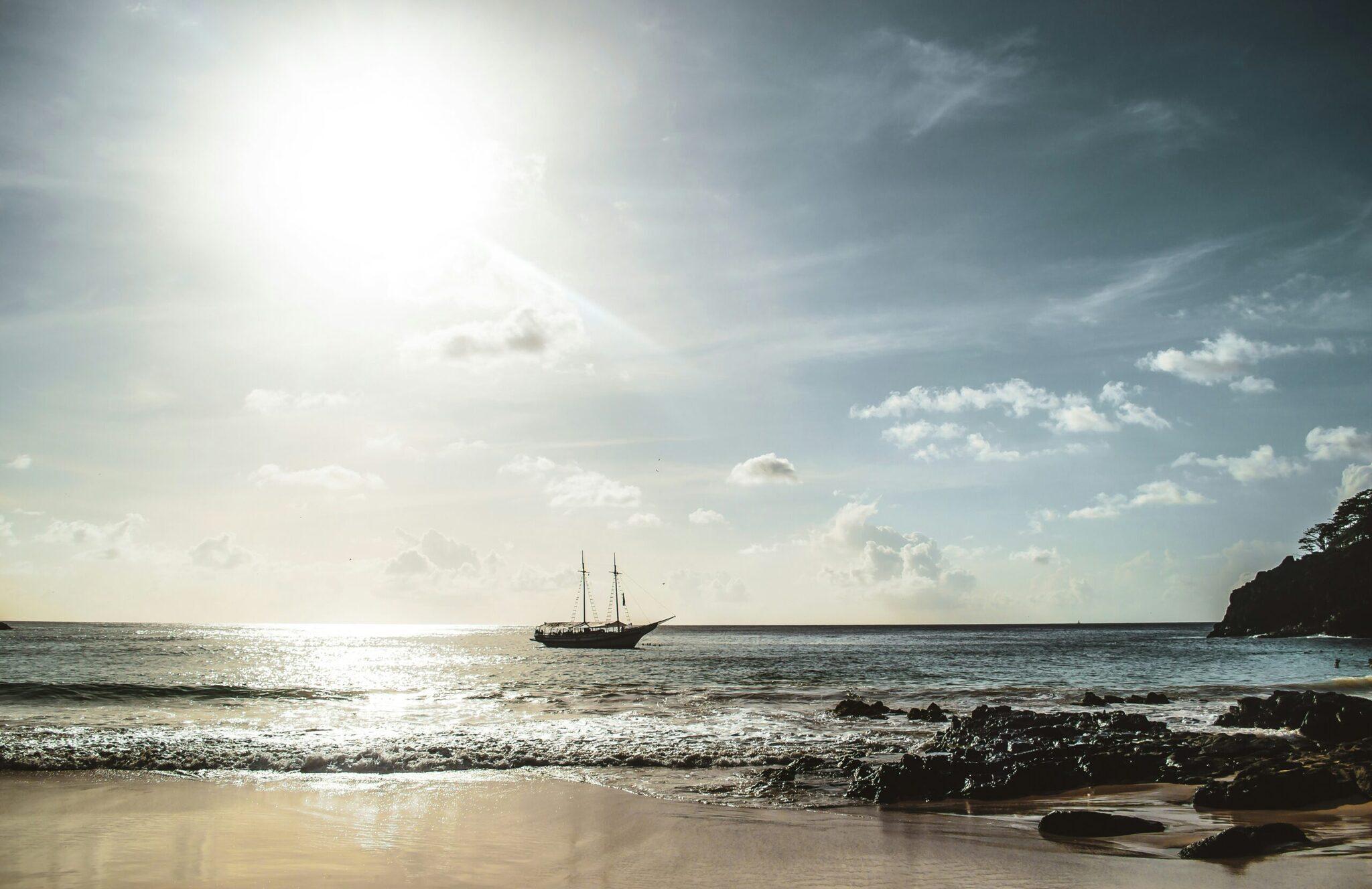 Un velero tradicional fondeado frente a una playa de arena fina bordeada de rocas bajo un cielo nublado con sol