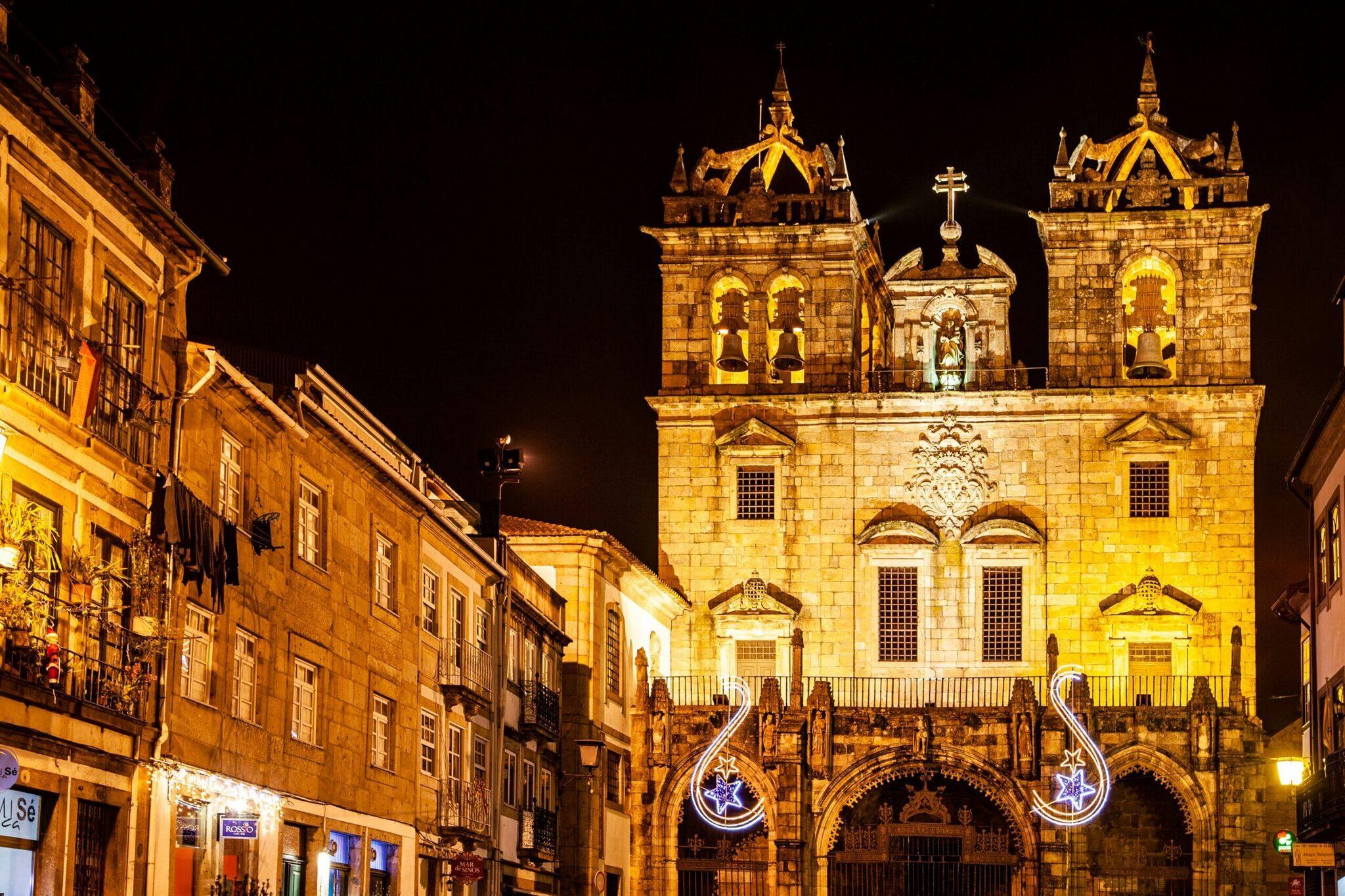 Fachada iluminada de una iglesia barroca con dos torres, decorada con motivos festivos luminosos, flanqueada por edificios históricos de piedra