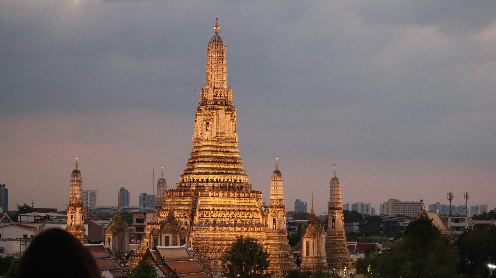 A grand gilded Thai Buddhist temple illuminated at sunset, set against the urban skyline