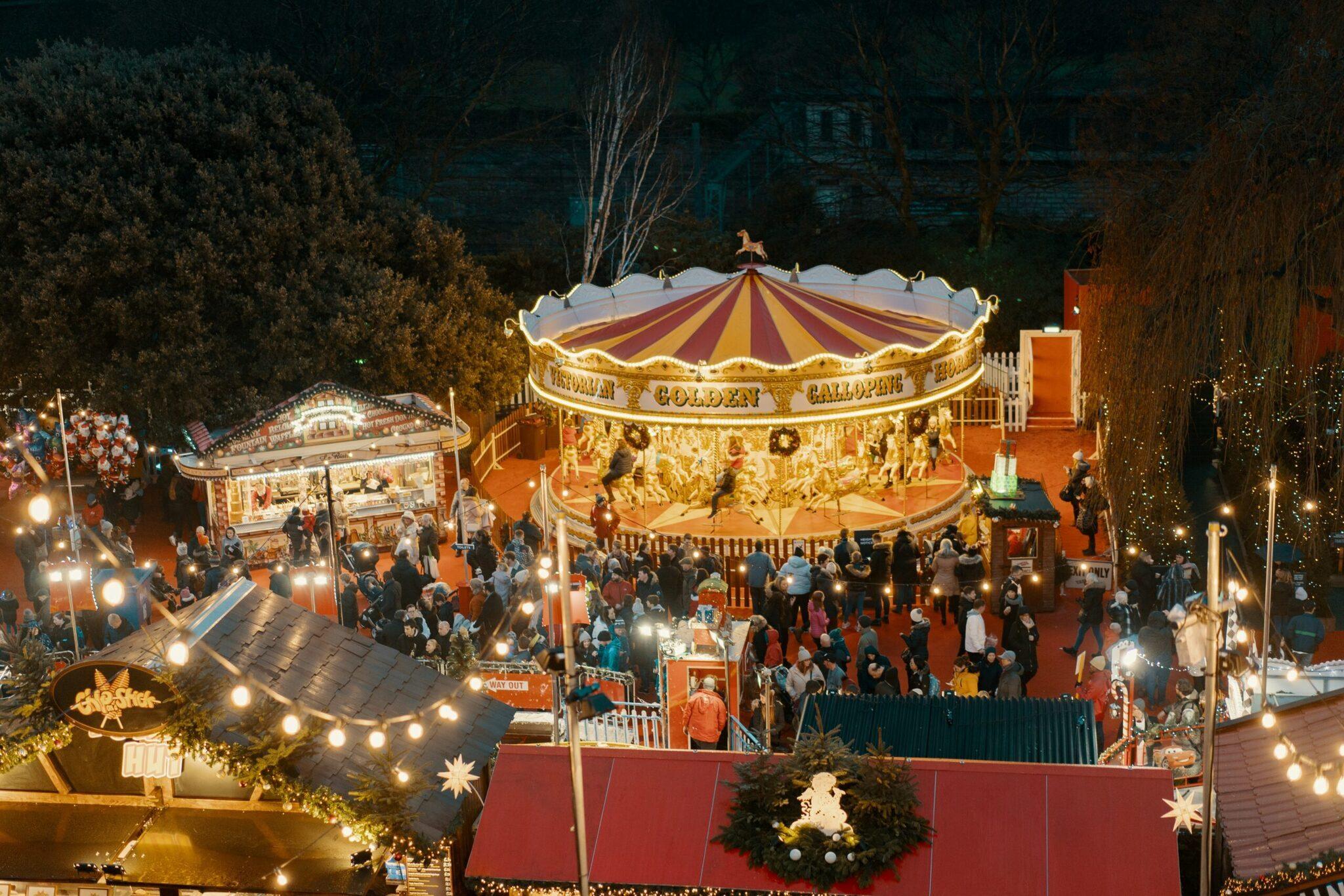 Mercado navideño nocturno con un tiovivo dorado iluminado y puestos decorados, visitantes bajo guirnaldas de luces