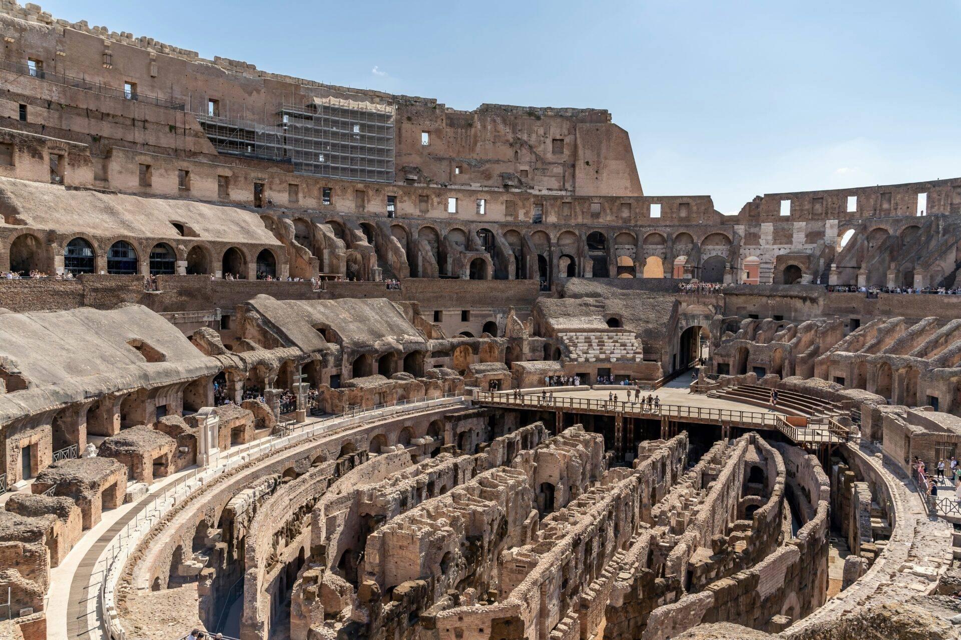 Interior of a ruined Roman amphitheatre showing the arena floor, collapsed seating tiers and well-preserved arched arcades