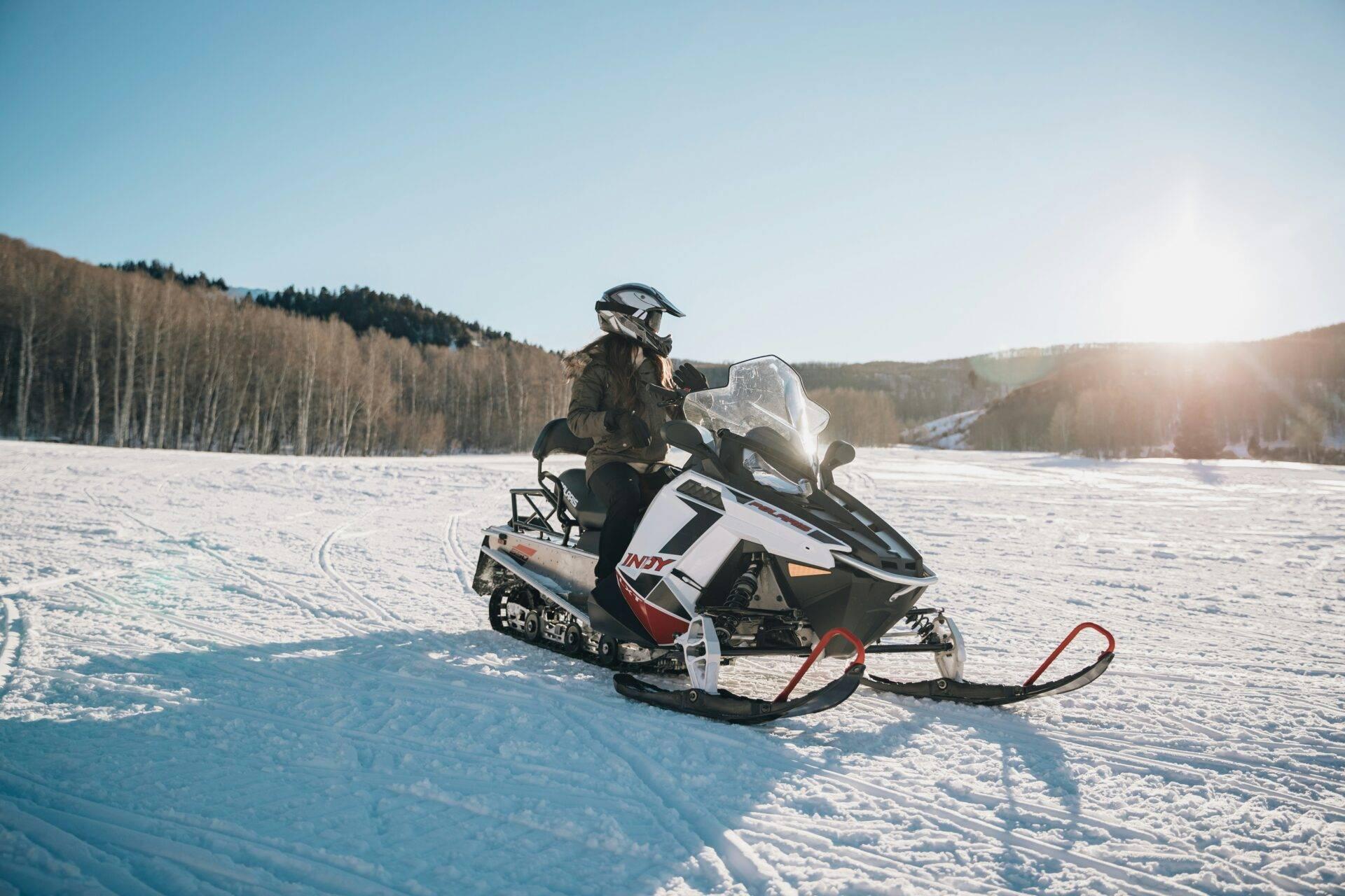 Ein Schneemobilfahrer in Winterausrüstung überquert einen zugefrorenen See umgeben von verschneiten Nadelwäldern