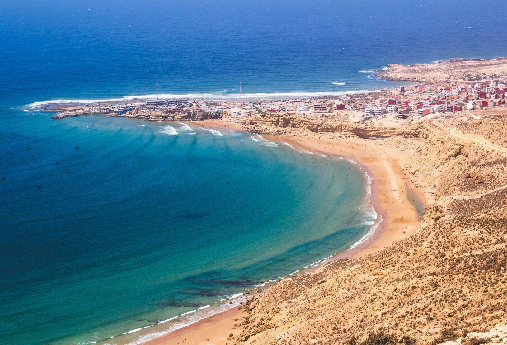 Luftaufnahme einer marokkanischen Küstenbucht mit Sandstrand, bebautem Hinterland und kristallklarem türkisfarbenem Wasser