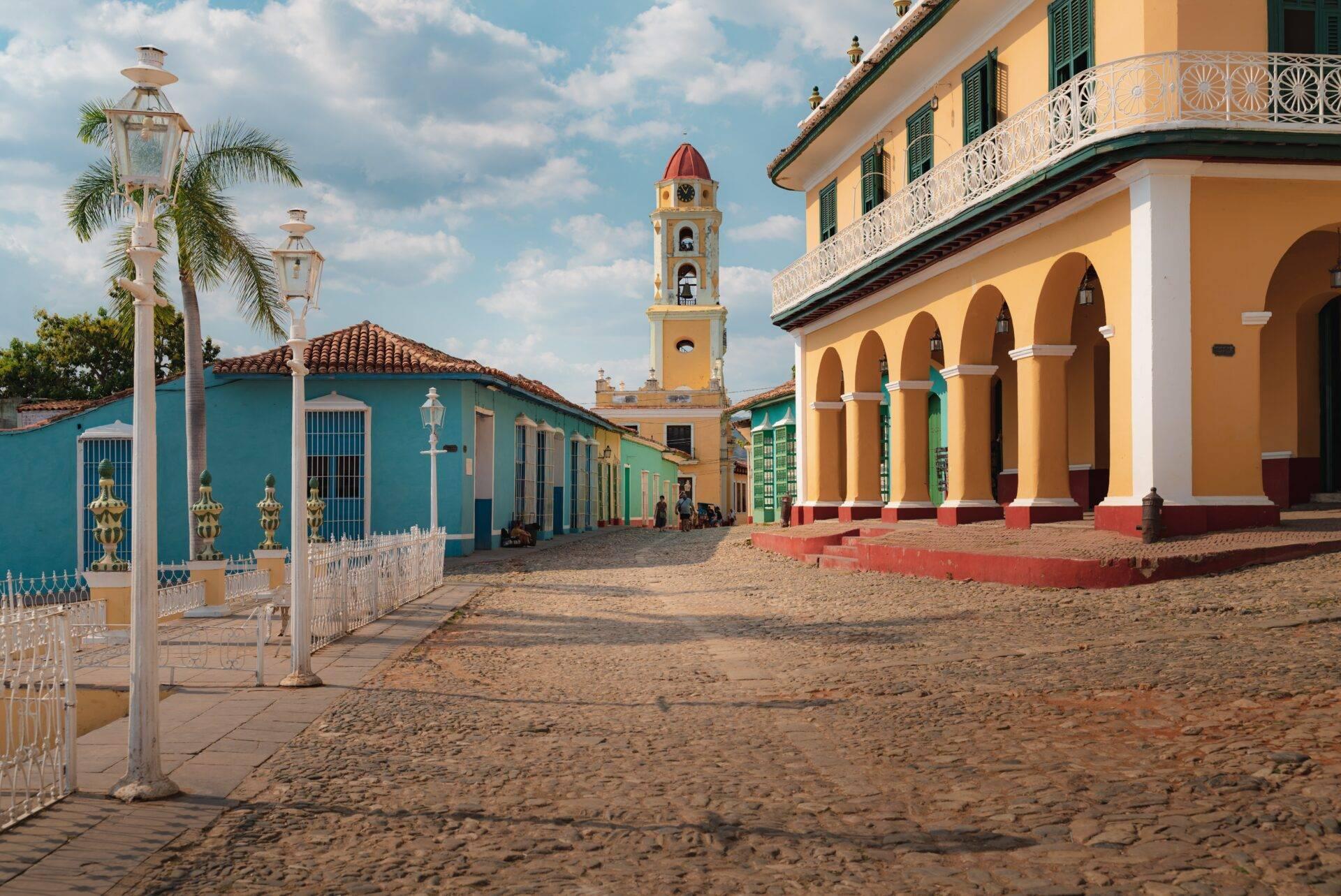 Una piazza coloniale colorata con edifici gialli e blu, torre con orologio rosso e arcate, sotto un cielo terso
