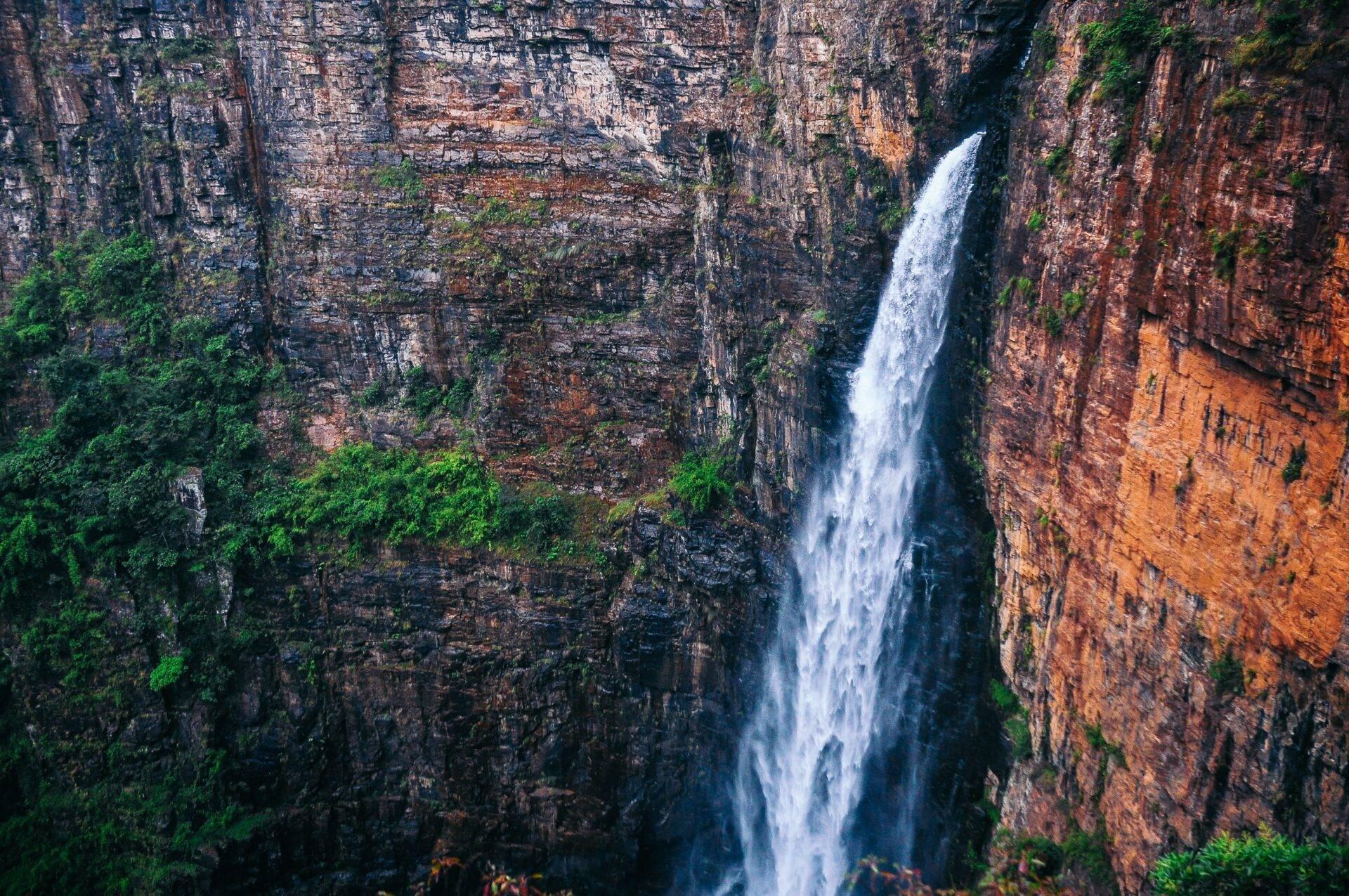 Una cascada blanca desciende por un acantilado de roca rojiza encajonado, cubierto de exuberante vegetación verde