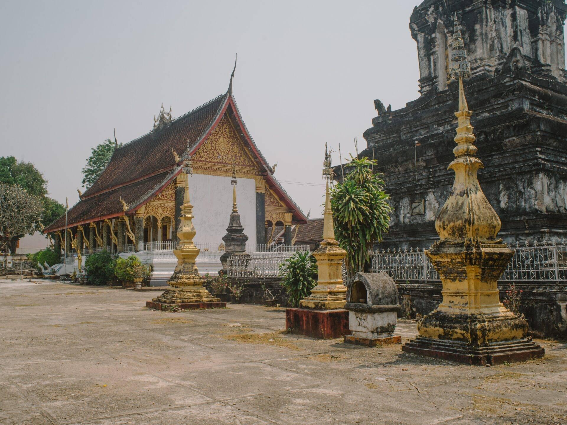 Cortile di un tempio buddista laotiano con stupa dorati, edificio principale dai tetti rossi e ornamenti dorati