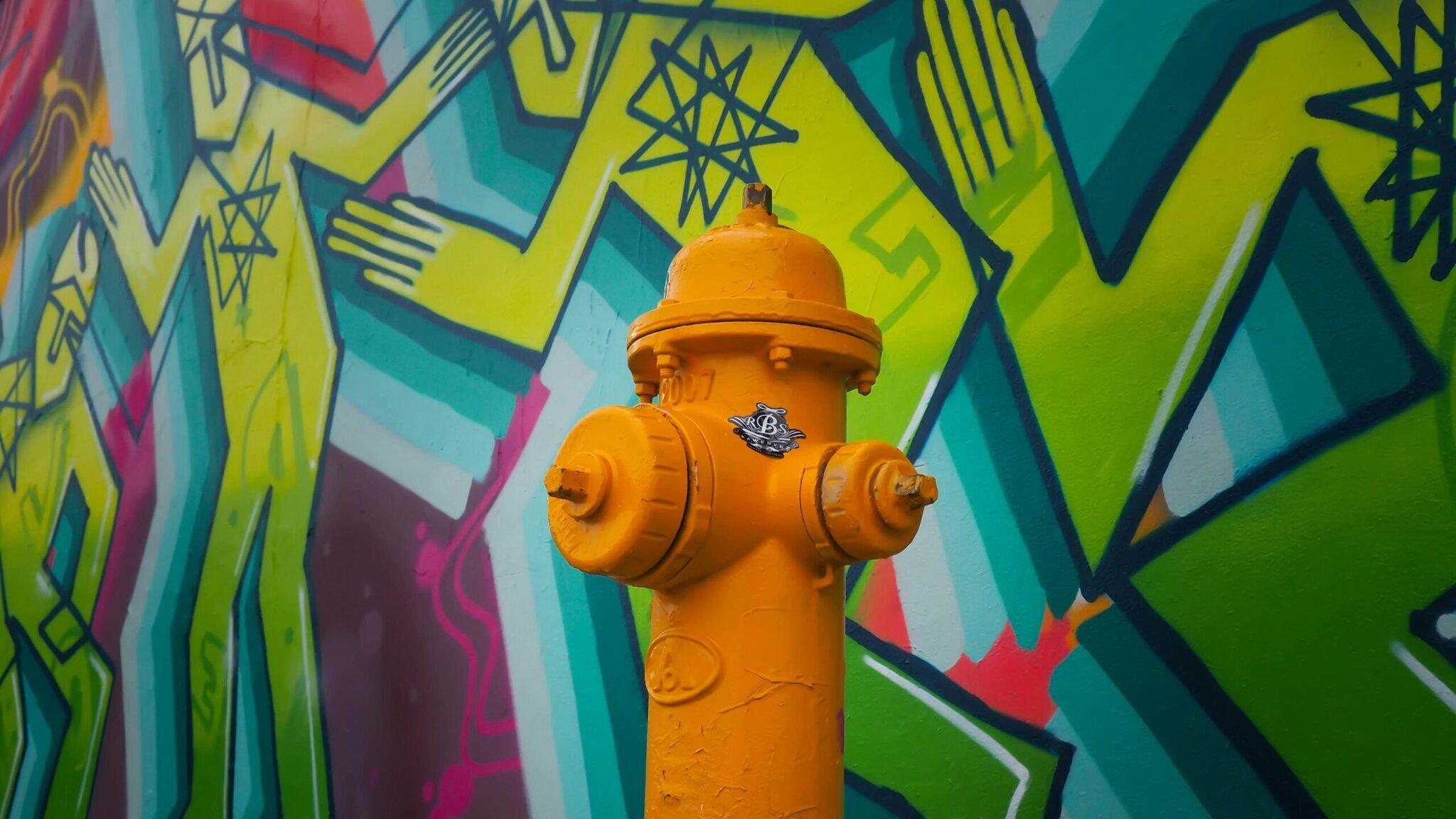 A bright orange fire hydrant in front of a vivid street art mural with geometric yellow and turquoise patterns