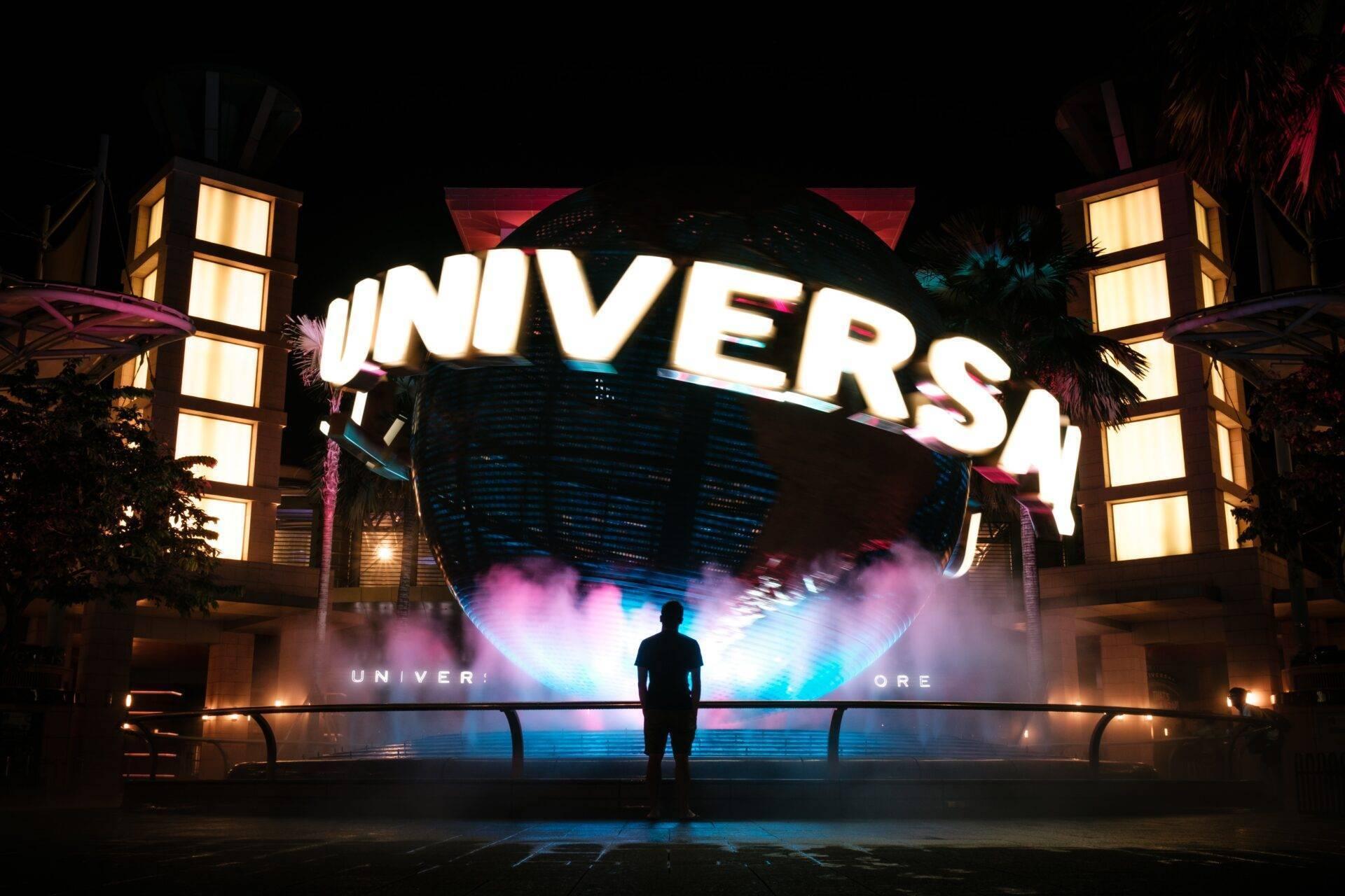 Ein Besucher vor dem beleuchteten Eingang von Universal Studios Singapore bei Nacht, mit riesigem Globus und farbigen Lichteffekten