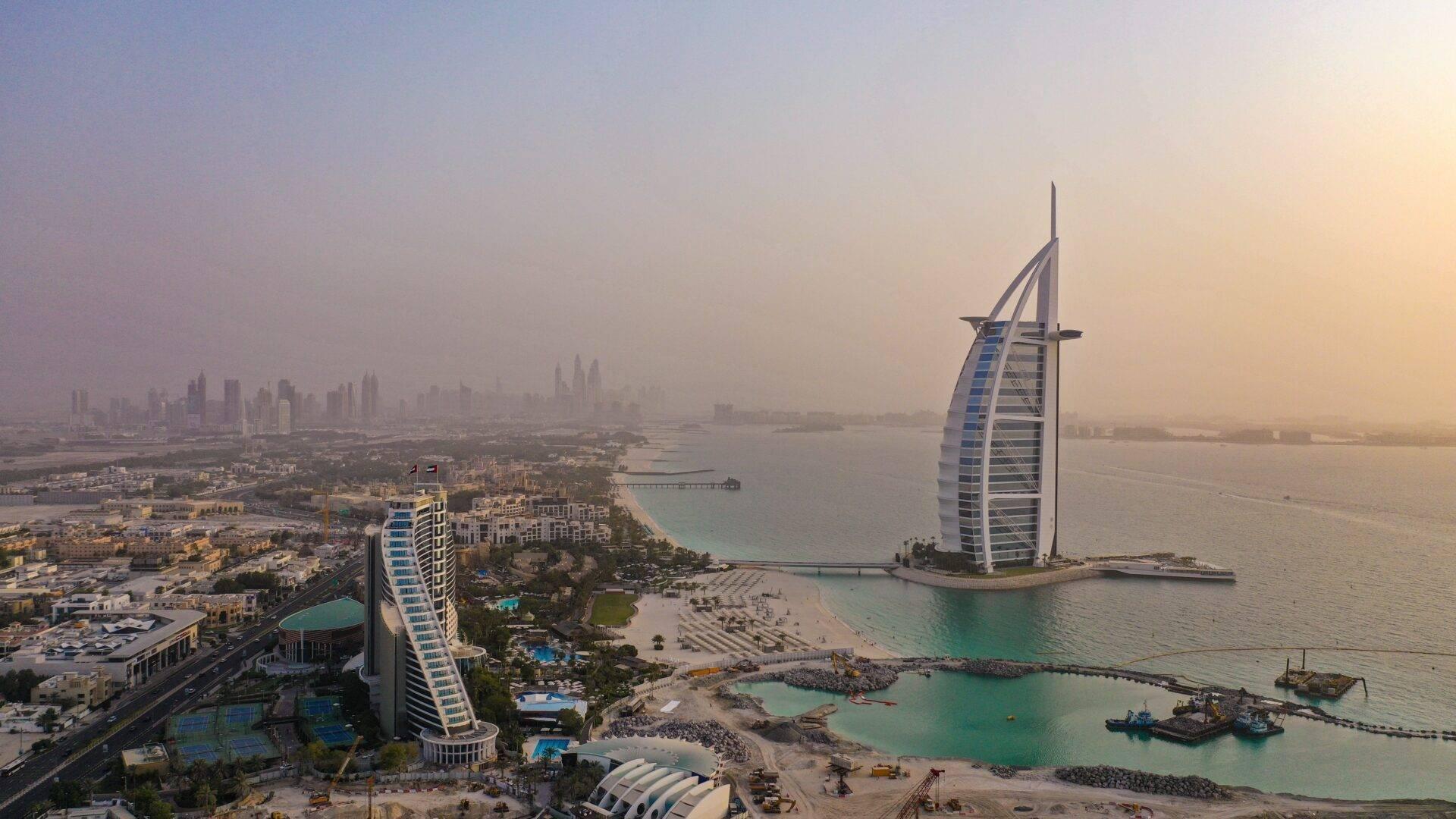 Luftaufnahme einer Küstenbucht mit zwei modernen segelförmigen Hotels, türkisfarbenen Stränden und urbaner Skyline bei Sonnenuntergang
