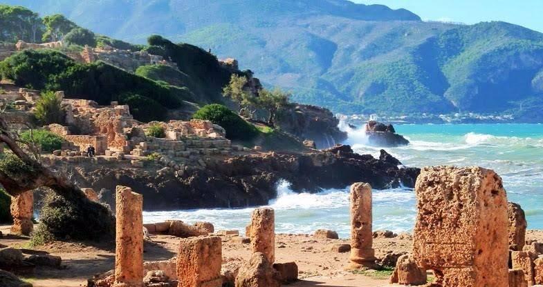 Ruinas antiguas con columnas de piedra ante una playa mediterránea de aguas turquesas y montañas verdes