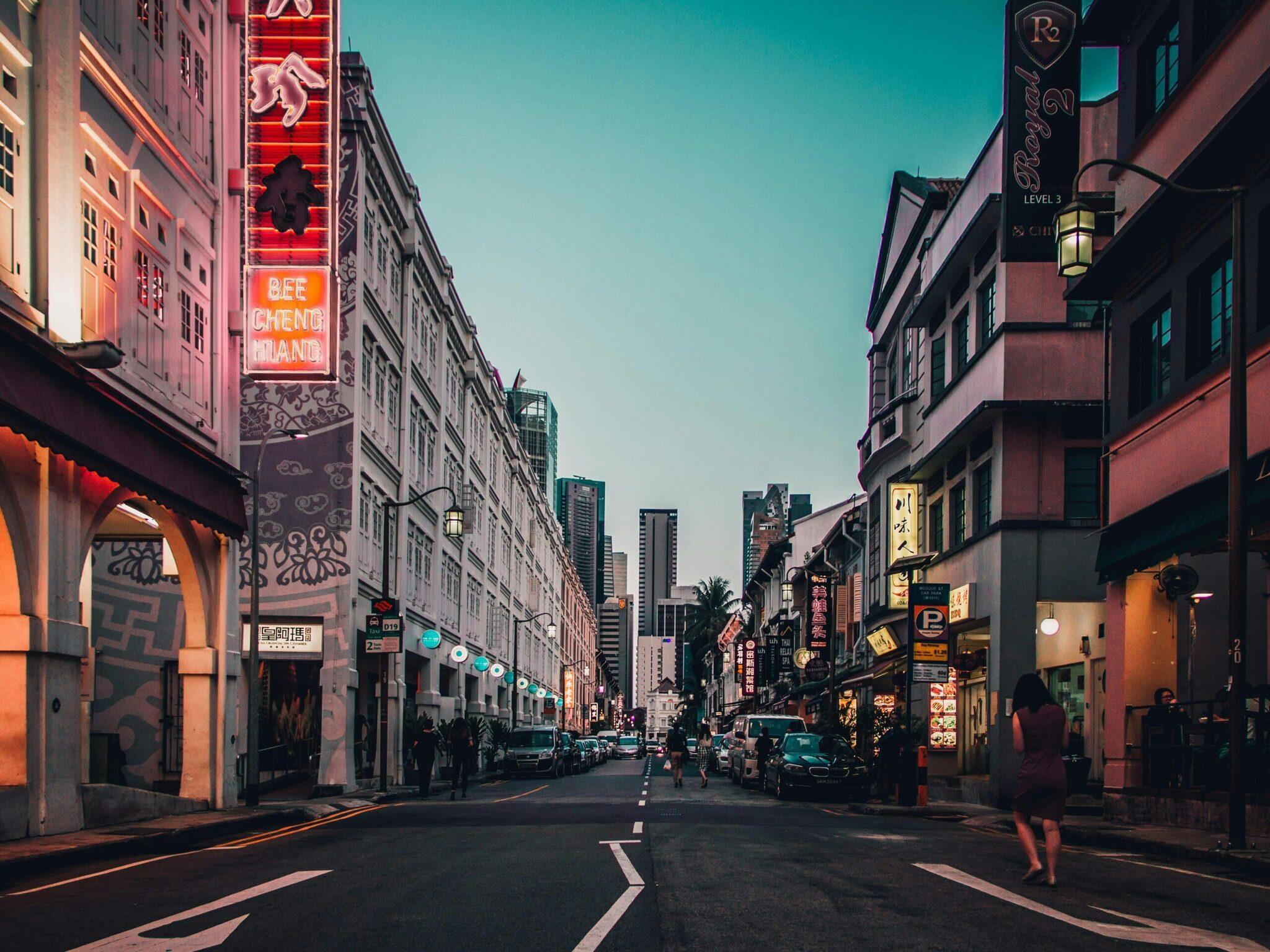 Una strada commerciale asiatica illuminata da neon rossi con palazzi storici e grattacieli sullo sfondo al crepuscolo
