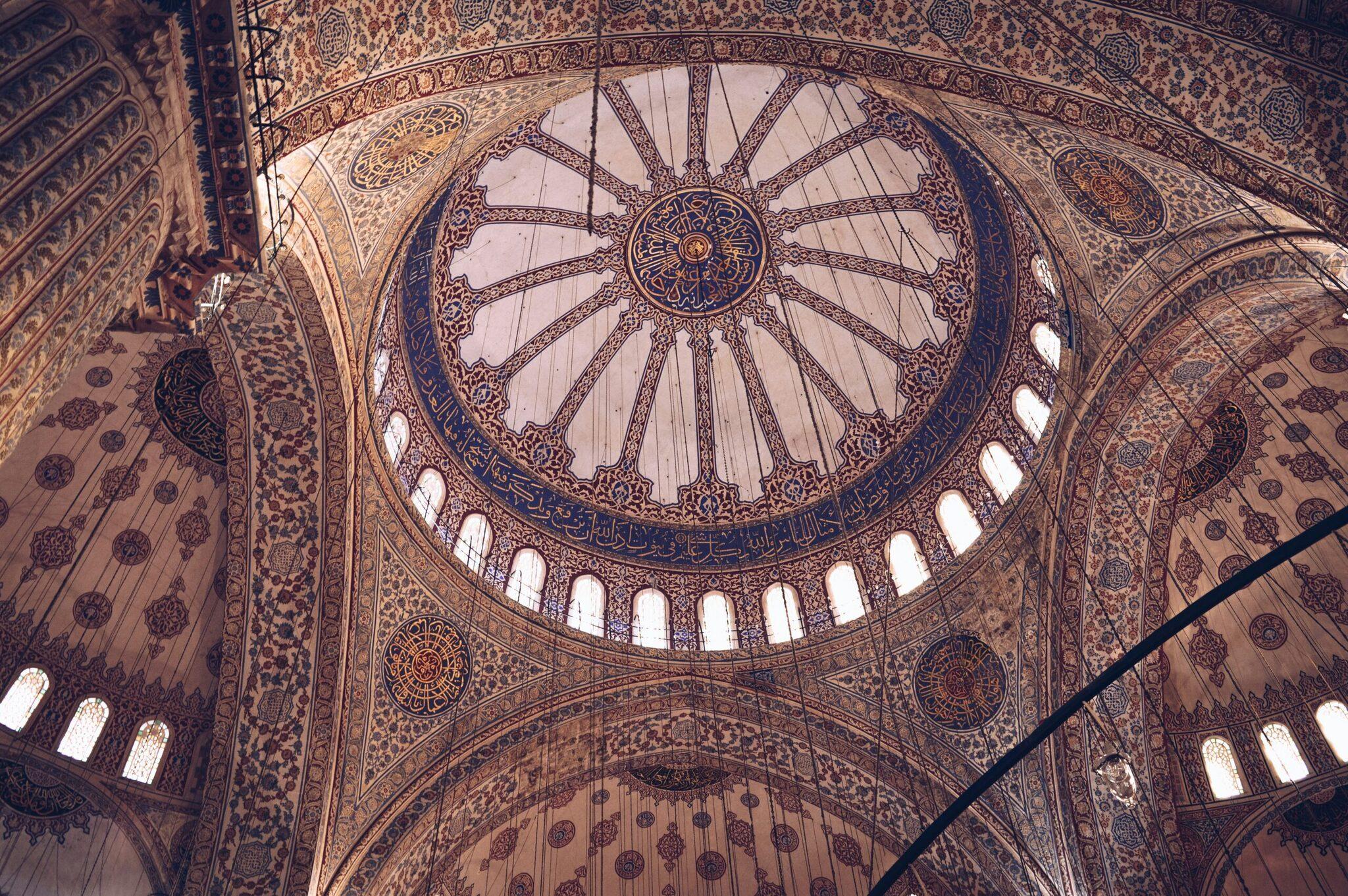 Interno di una moschea con cupola centrale ornata da motivi geometrici e calligrafia islamica, luce naturale dalle finestre