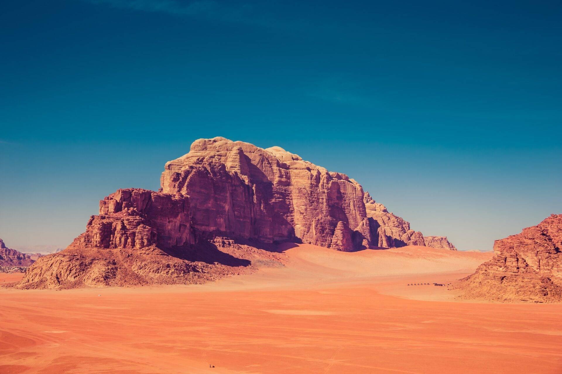 Un macizo rocoso rojo ocre se alza en un desierto de arena rojiza bajo un cielo azul despejado