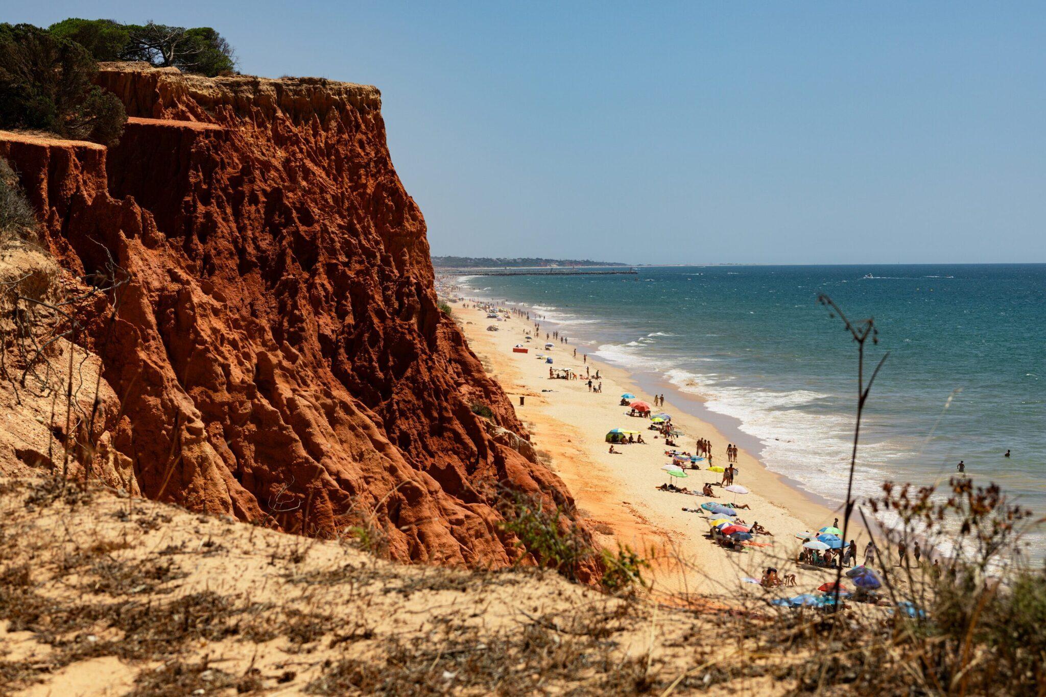 Ochre cliff towering above a golden sandy beach with swimmers and parasols under a clear blue sky