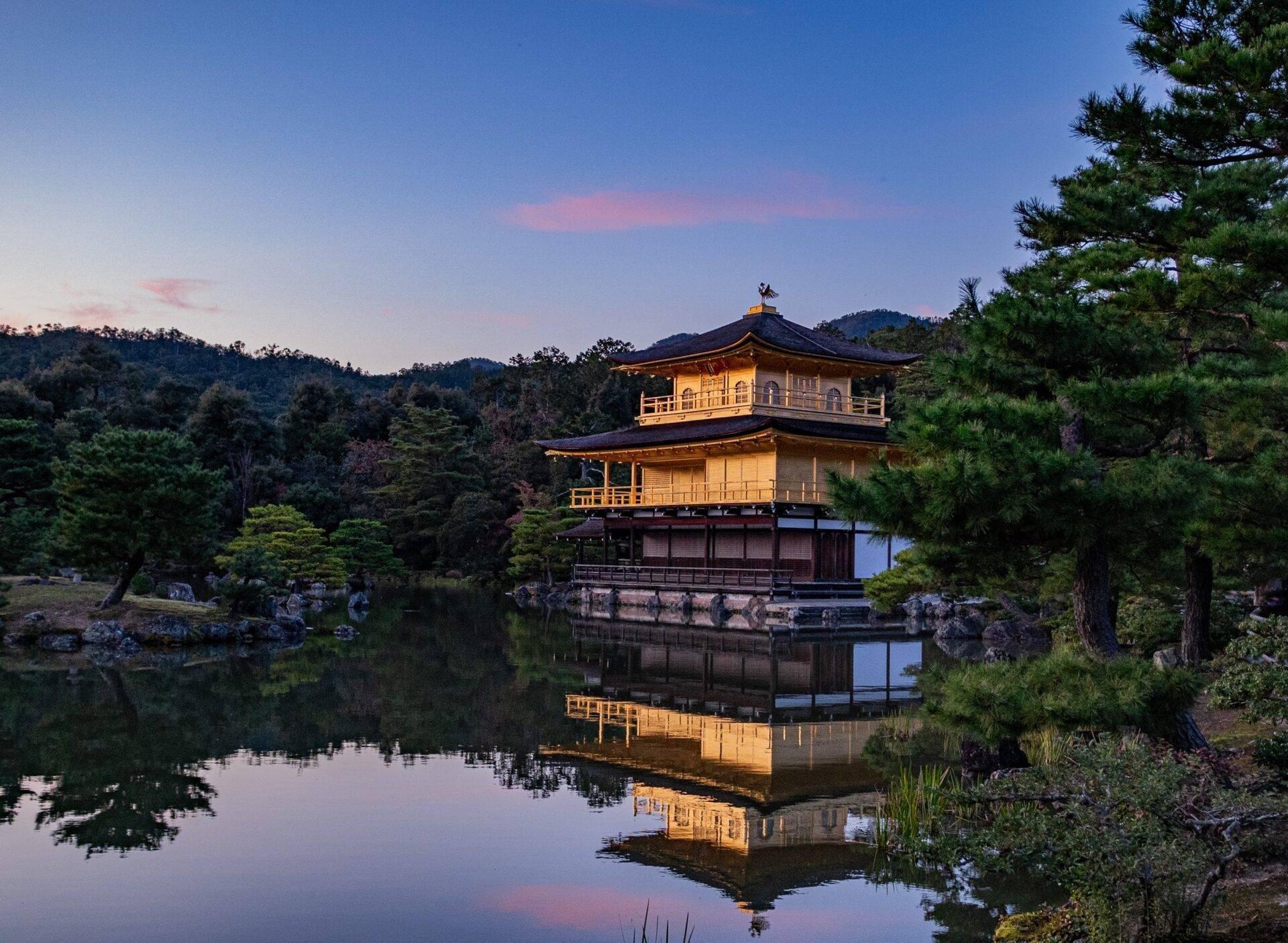 Ein zweistöckiger goldener Pavillon spiegelt sich in einem ruhigen See, umgeben von üppigen Wäldern in der Abenddämmerung