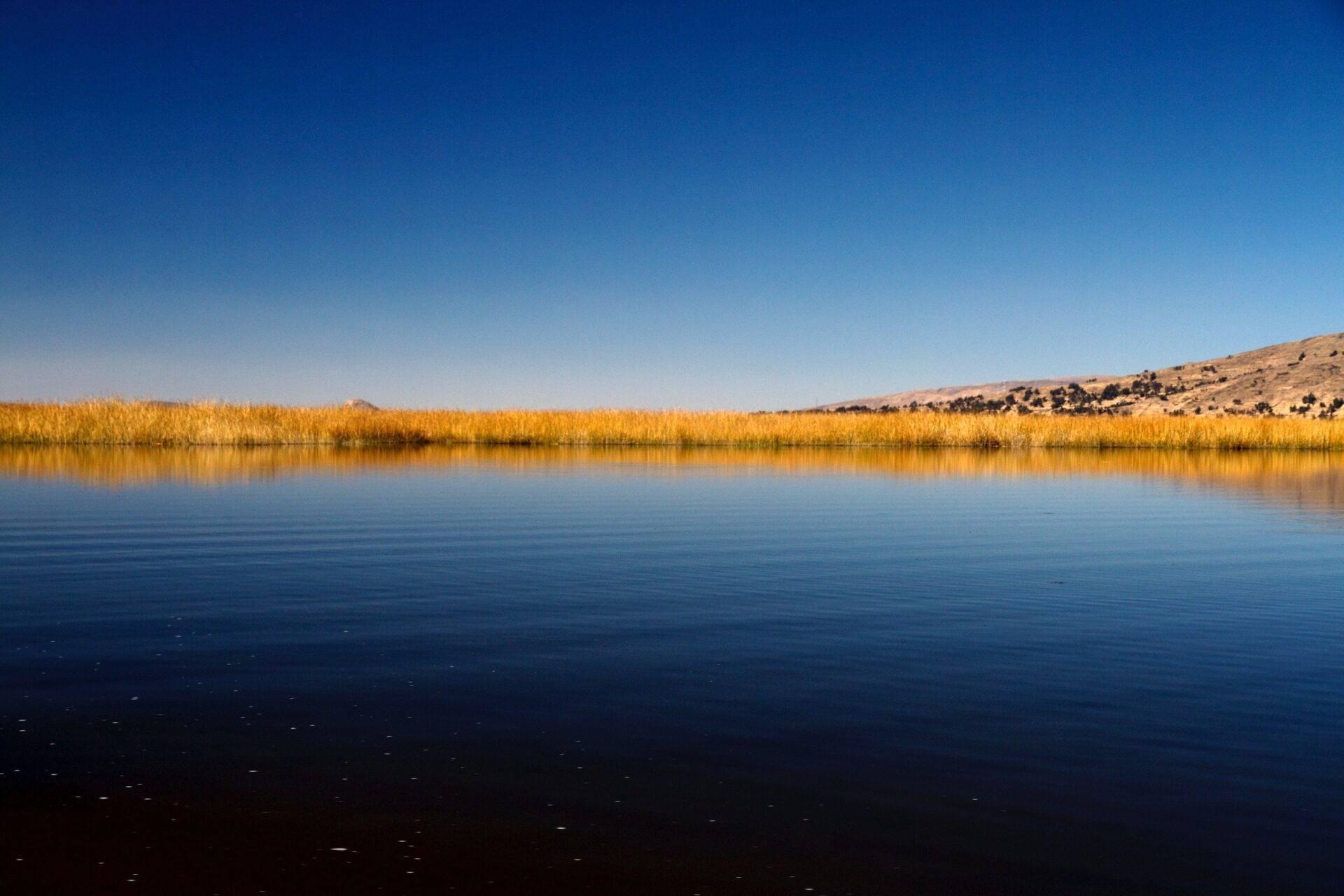 Lago calmo dalle acque turchesi circondato da canne dorate e montagne aride sotto un cielo azzurro terso