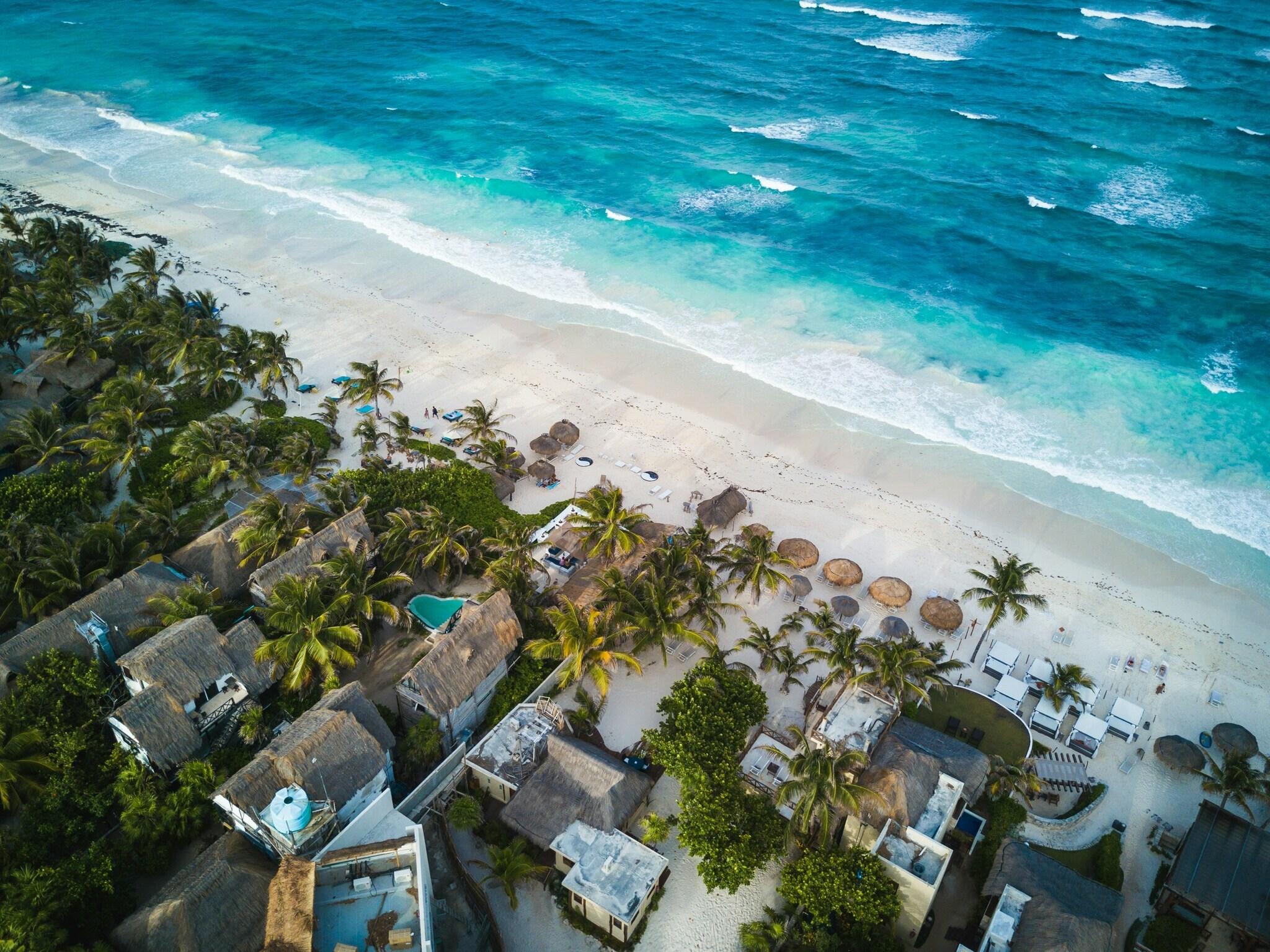 Luftaufnahme eines Tropenstrandes mit Resort, Palmen, Strohdächern und türkisfarbenem Wasser