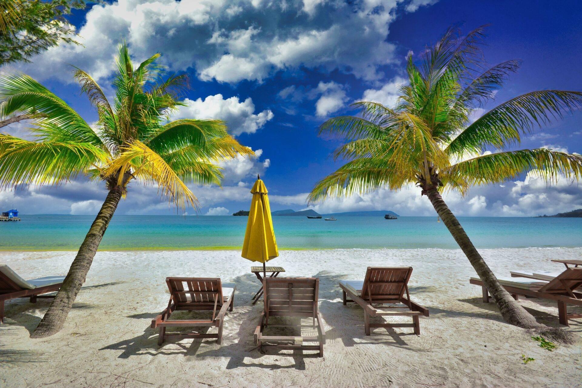 Tropischer Strand mit Holzliegen, gelbem Sonnenschirm und Palmen, umrahmt von türkisblauem Meer unter bewölktem blauen Himmel
