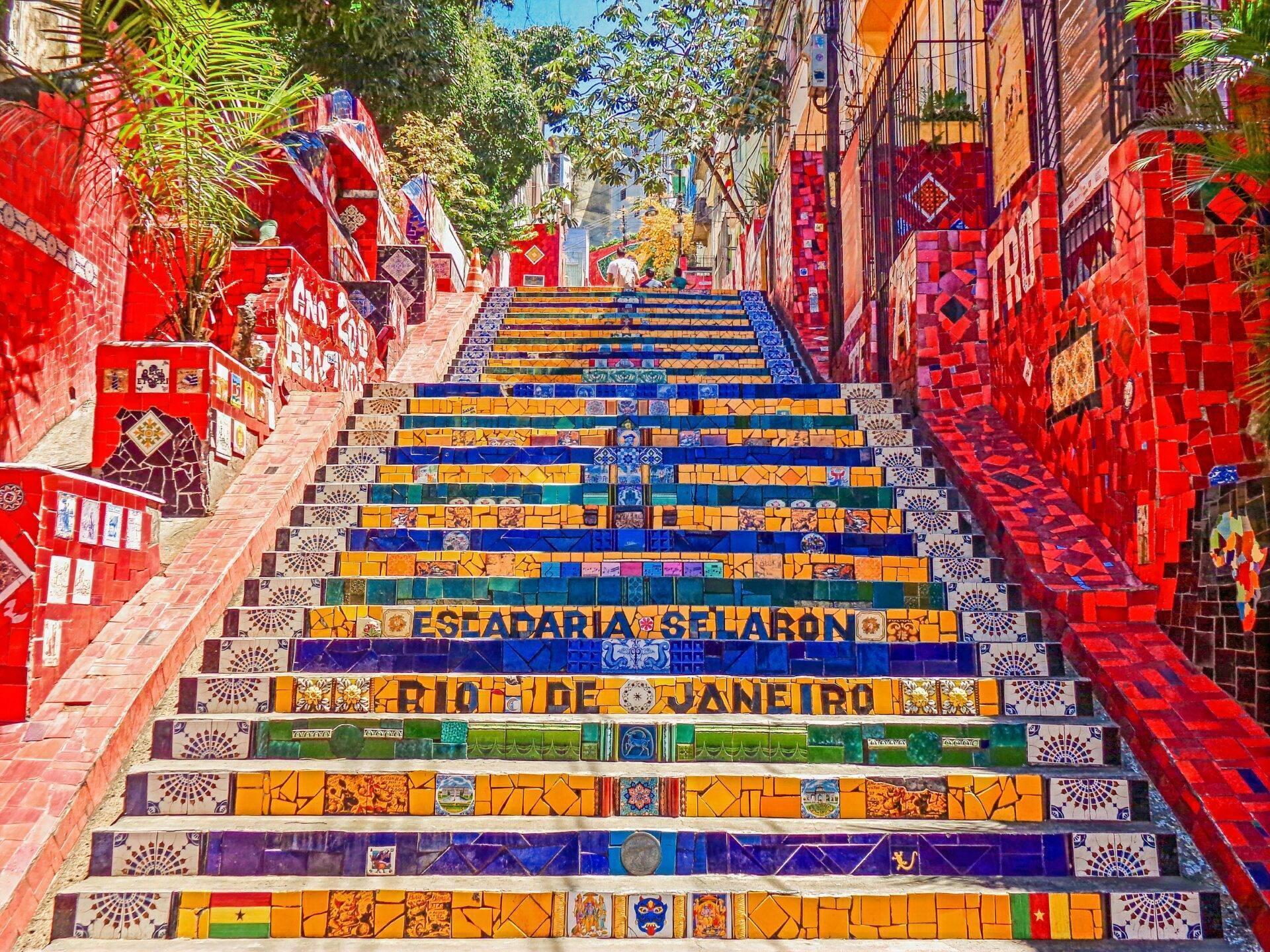 Scalinata monumentale dai gradini colorati decorati con ceramiche, circondata da muri rossi ornati, in un vicolo lastricato brasiliano