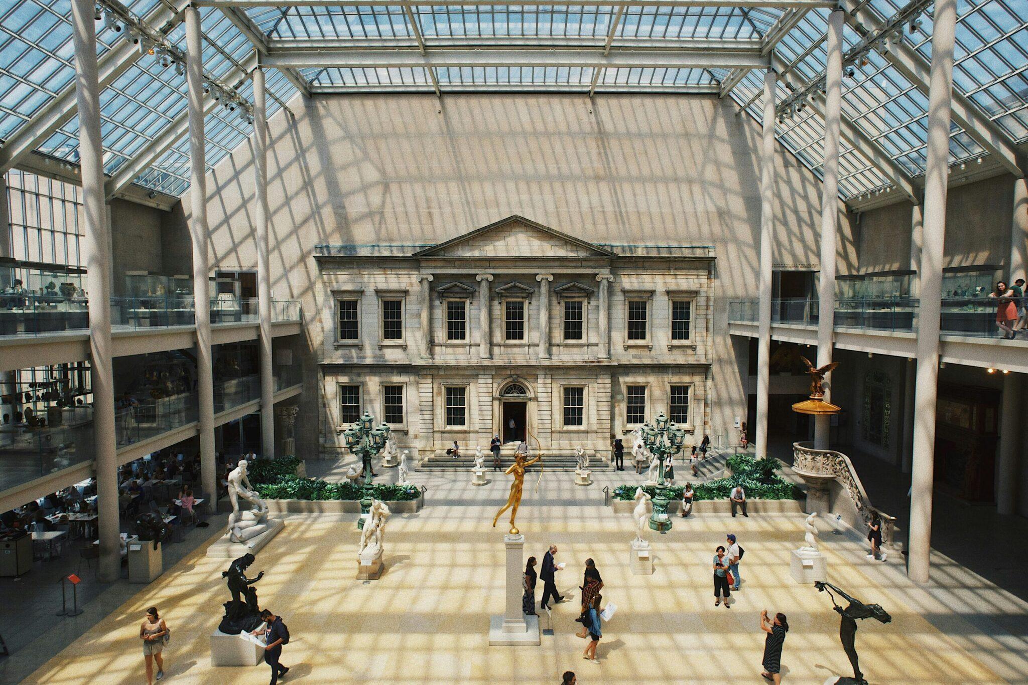 Vast museum interior with a central neoclassical building, sculptures and visitors beneath a contemporary glass roof