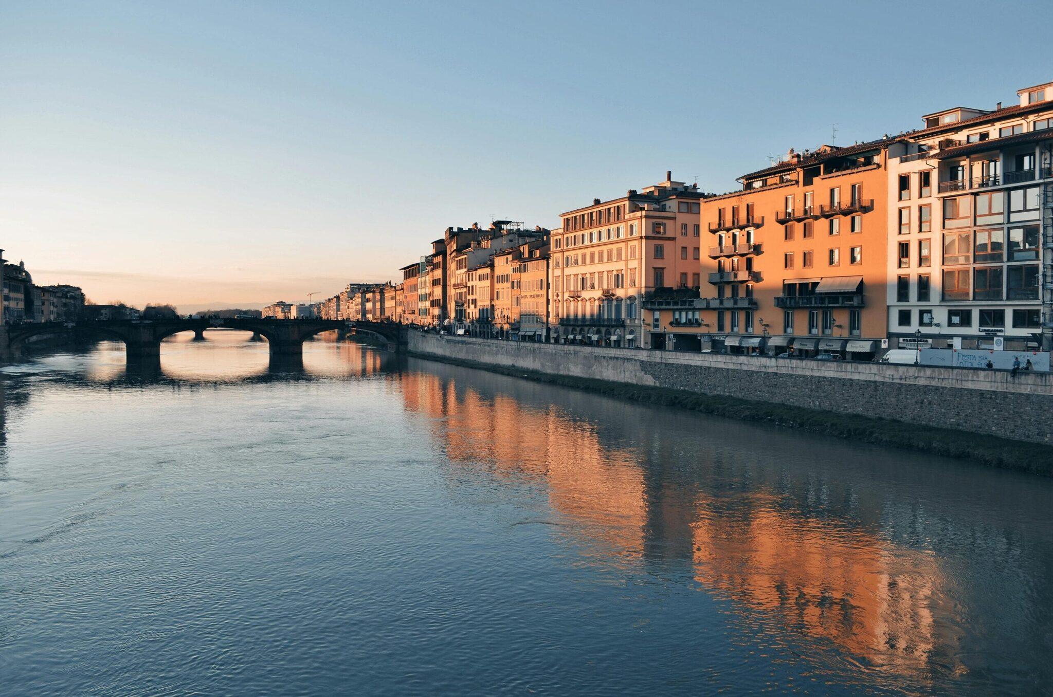 Edificios históricos de fachadas ocres reflejados en un río con un puente arqueado al atardecer