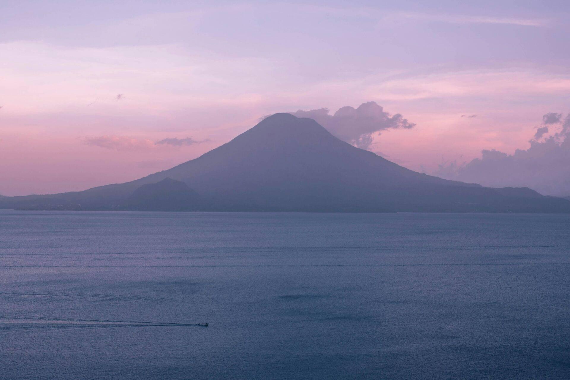 Vulkan mit regelmäßigen Hängen, der einen ruhigen See bei Sonnenuntergang in einer rosa-violetten Atmosphäre überragt