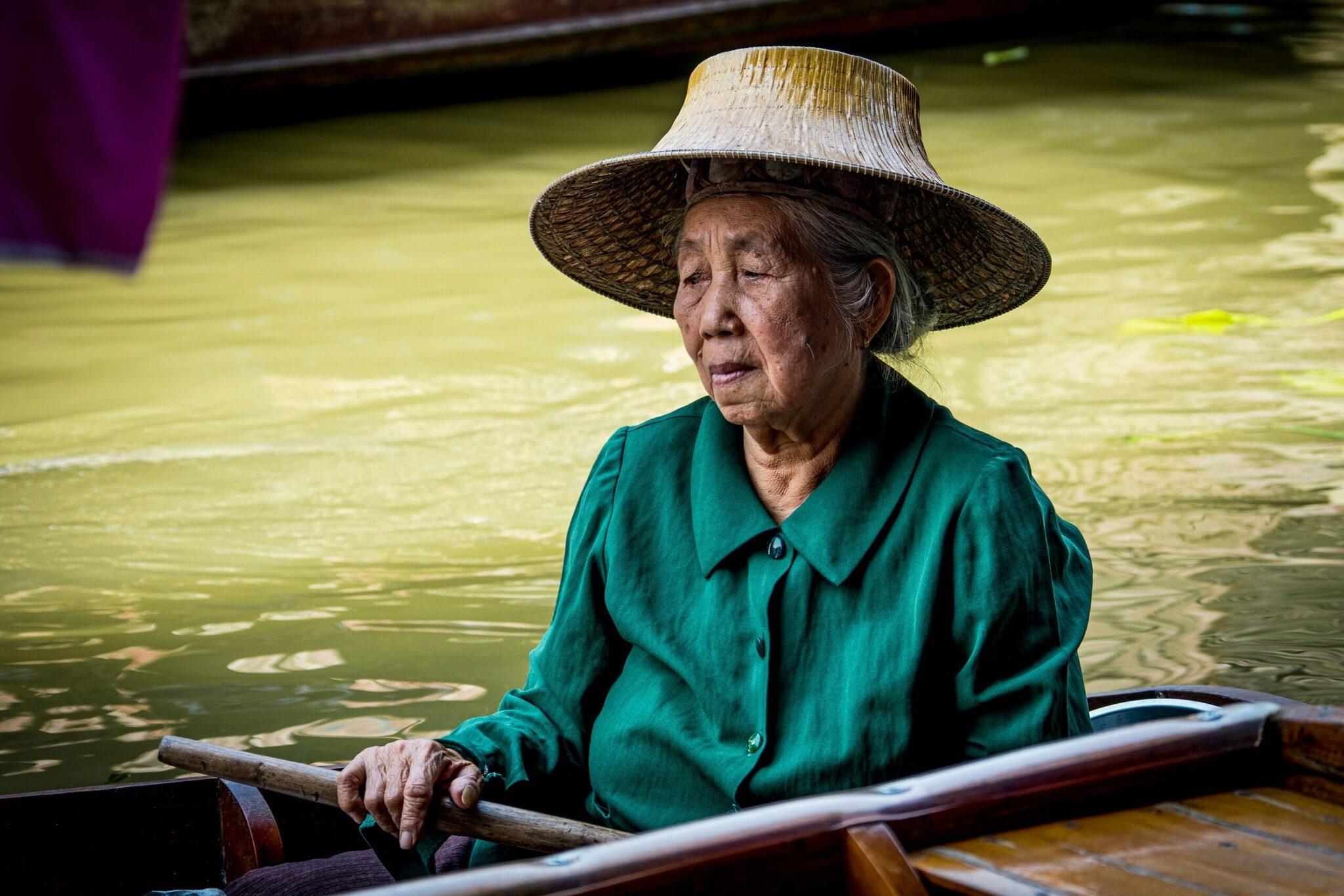 An elderly woman in a green shirt and traditional conical hat paddling a boat along a green canal